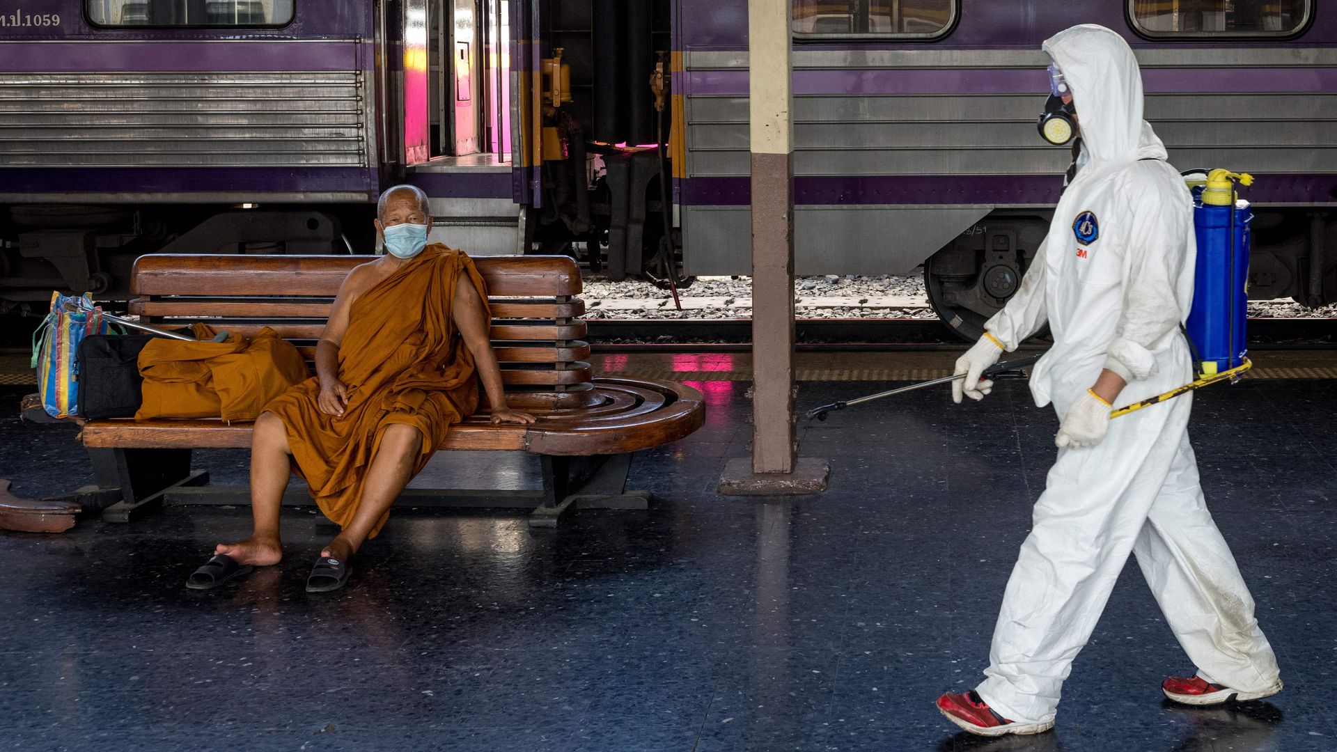 A cleaner wearing personal protective equipment (PPE) walks past a monk wearing a face mask at Hua Lamphong railway station in Bangkok on May 1