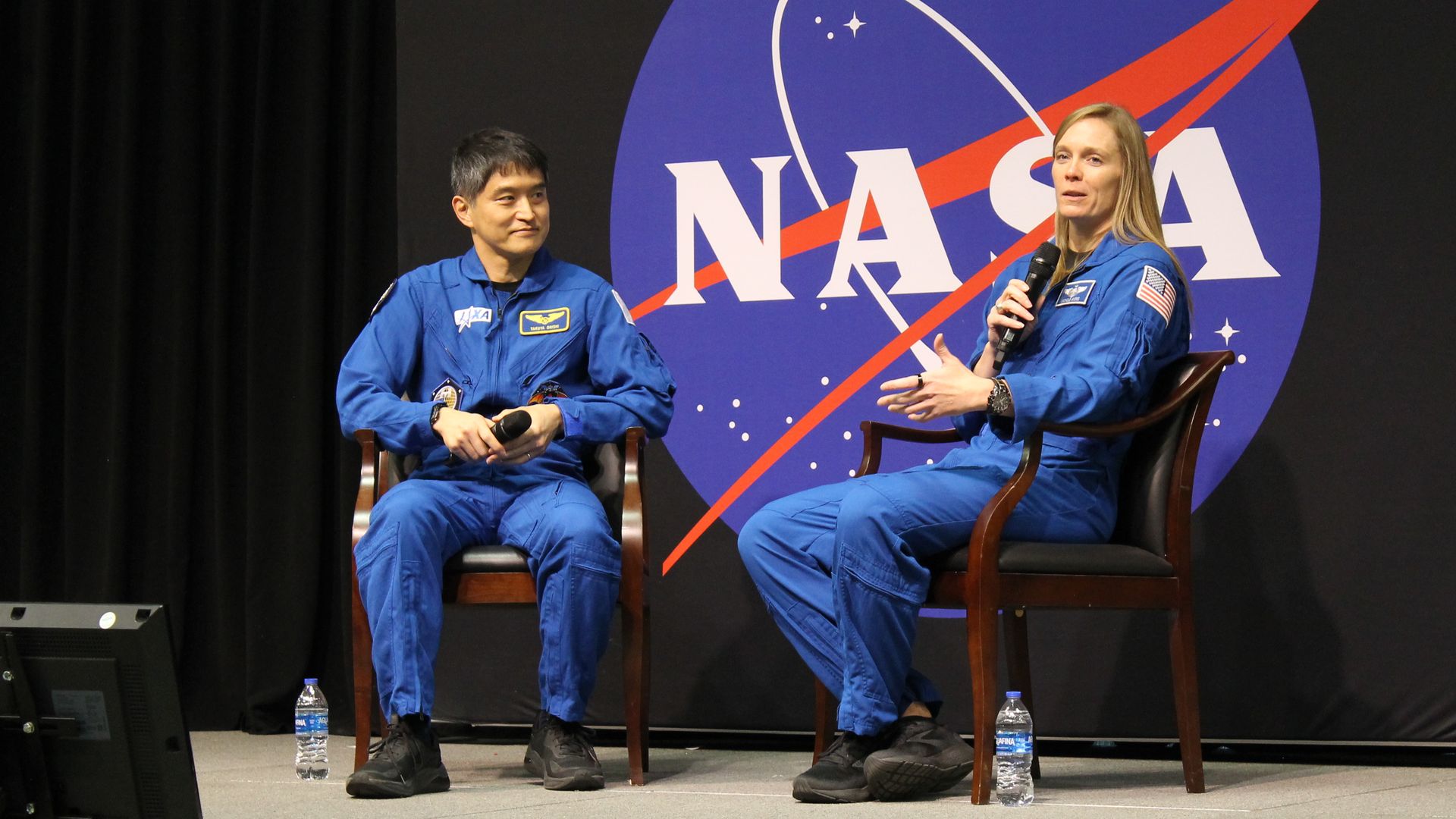 Two astronauts in blue jumpsuits holding microphones, sitting on chairs on stage with a large NASA logo backdrop. Water bottles on the floor beside each chair.