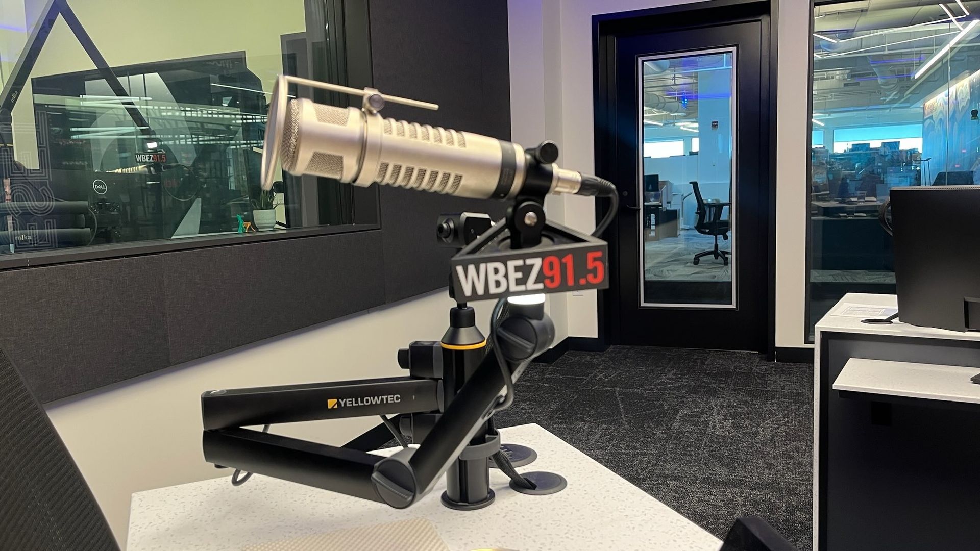 Close-up of a silver microphone with WBEZ 91.5 logo on a black Yellowtec arm in a radio studio with glass windows, black carpet, and visible office space behind a door and windows.