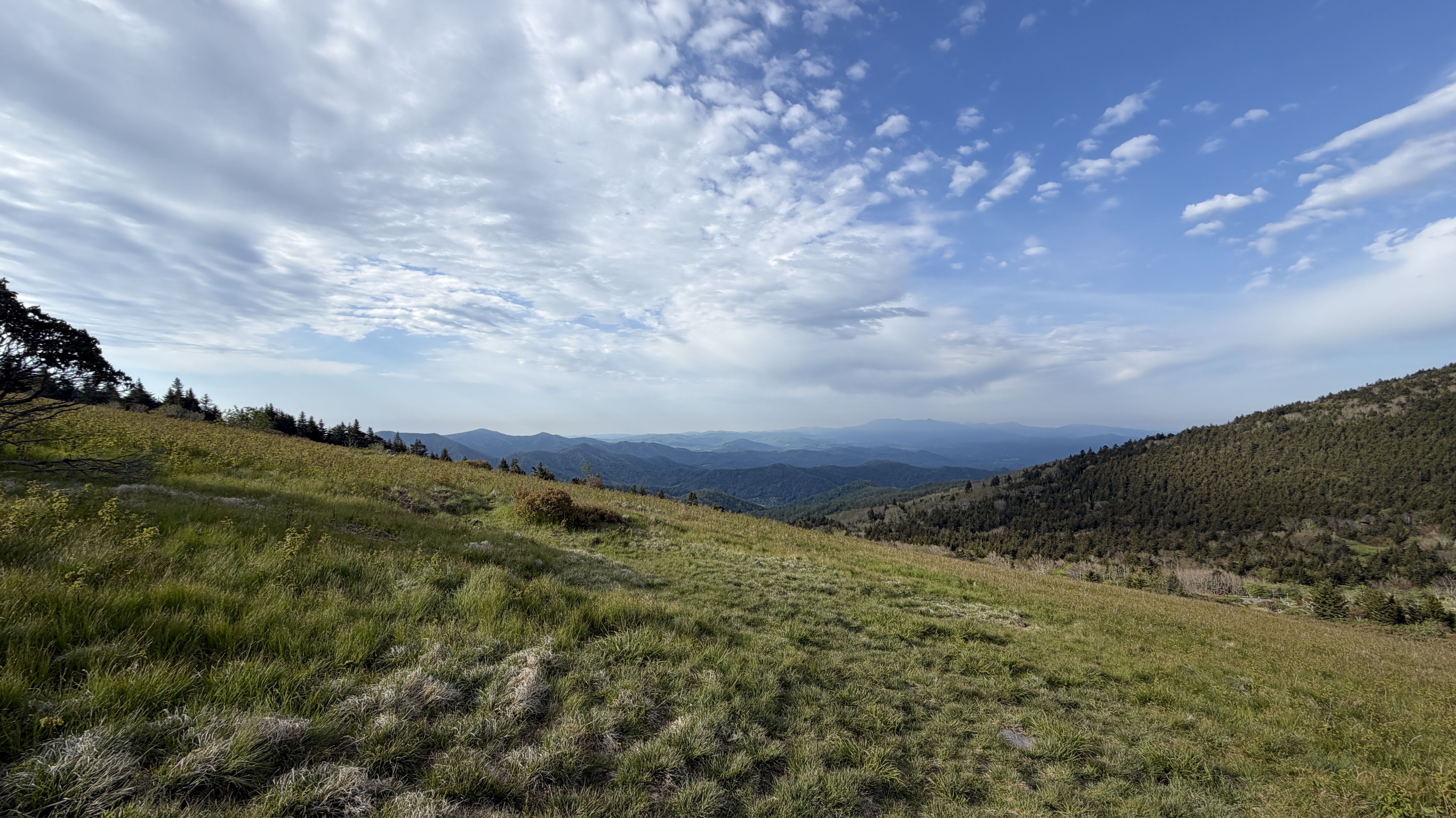 Wide hillside landscape with green grasses in the foreground, rolling hills and distant blue mountains under a partly cloudy blue sky.