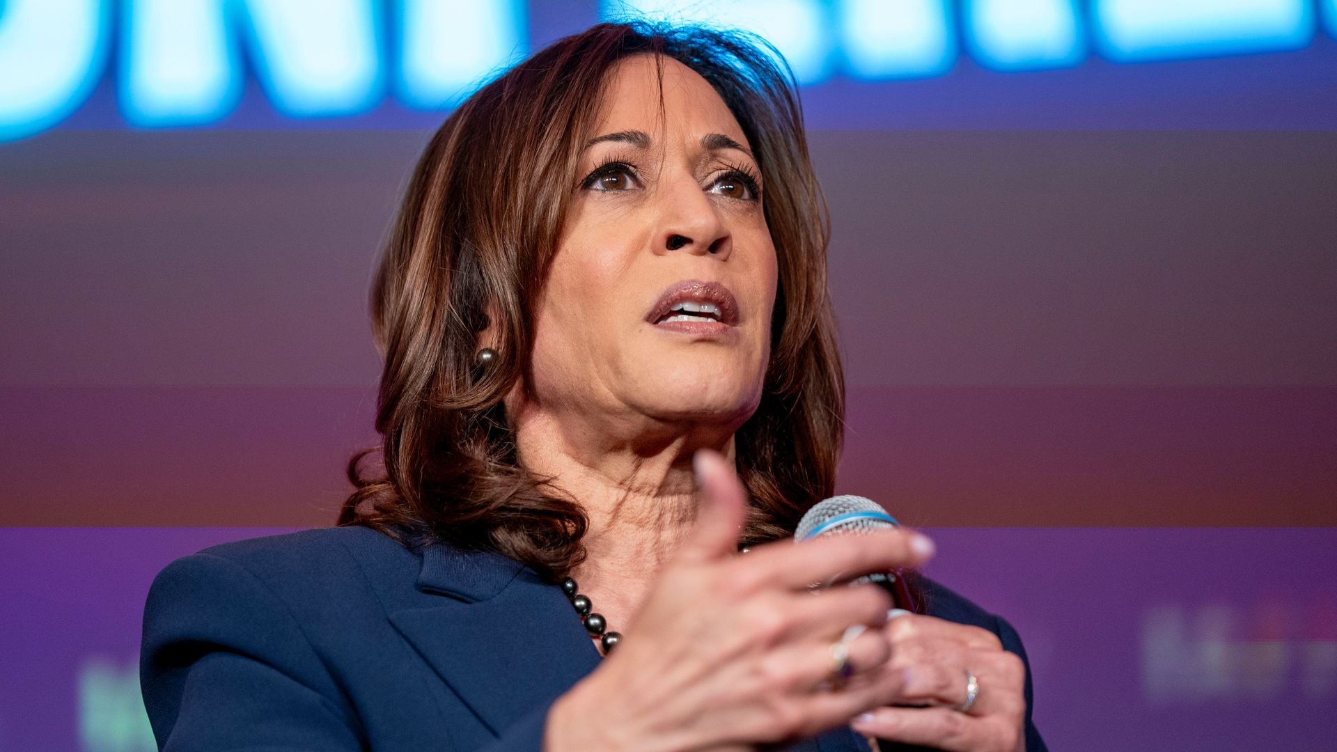 Vice President Kamala Harris, wearing a blue suit, holds a microphone as she speaks to an audience.