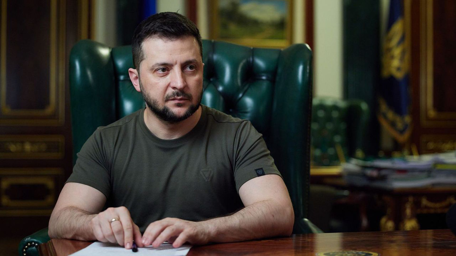 Photo of Volodymyr Zelenskyy sitting at a table with his hands resting on a piece of paper as he looks to his left