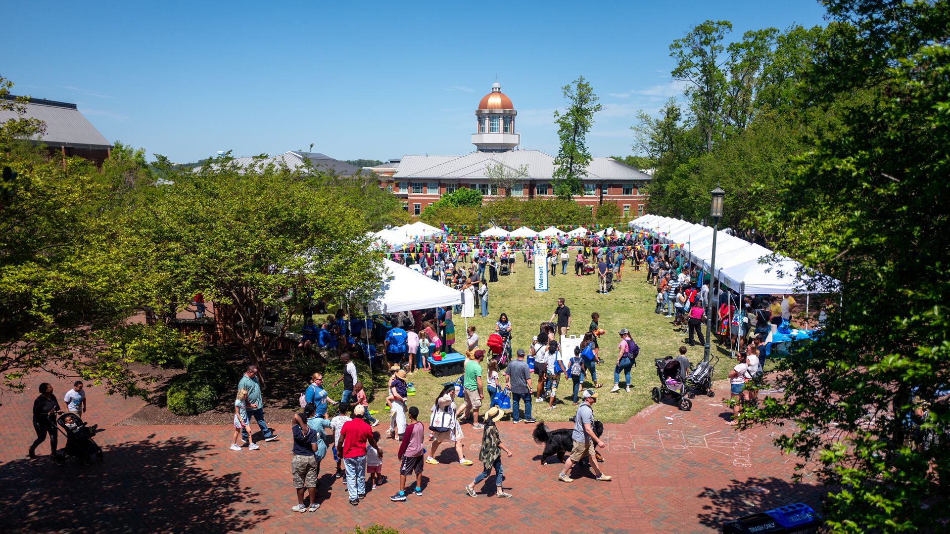 Outdoor community event with people walking around white tents and booths on a green lawn, colorful pennant banners overhead, and a large building with a copper dome in the background.