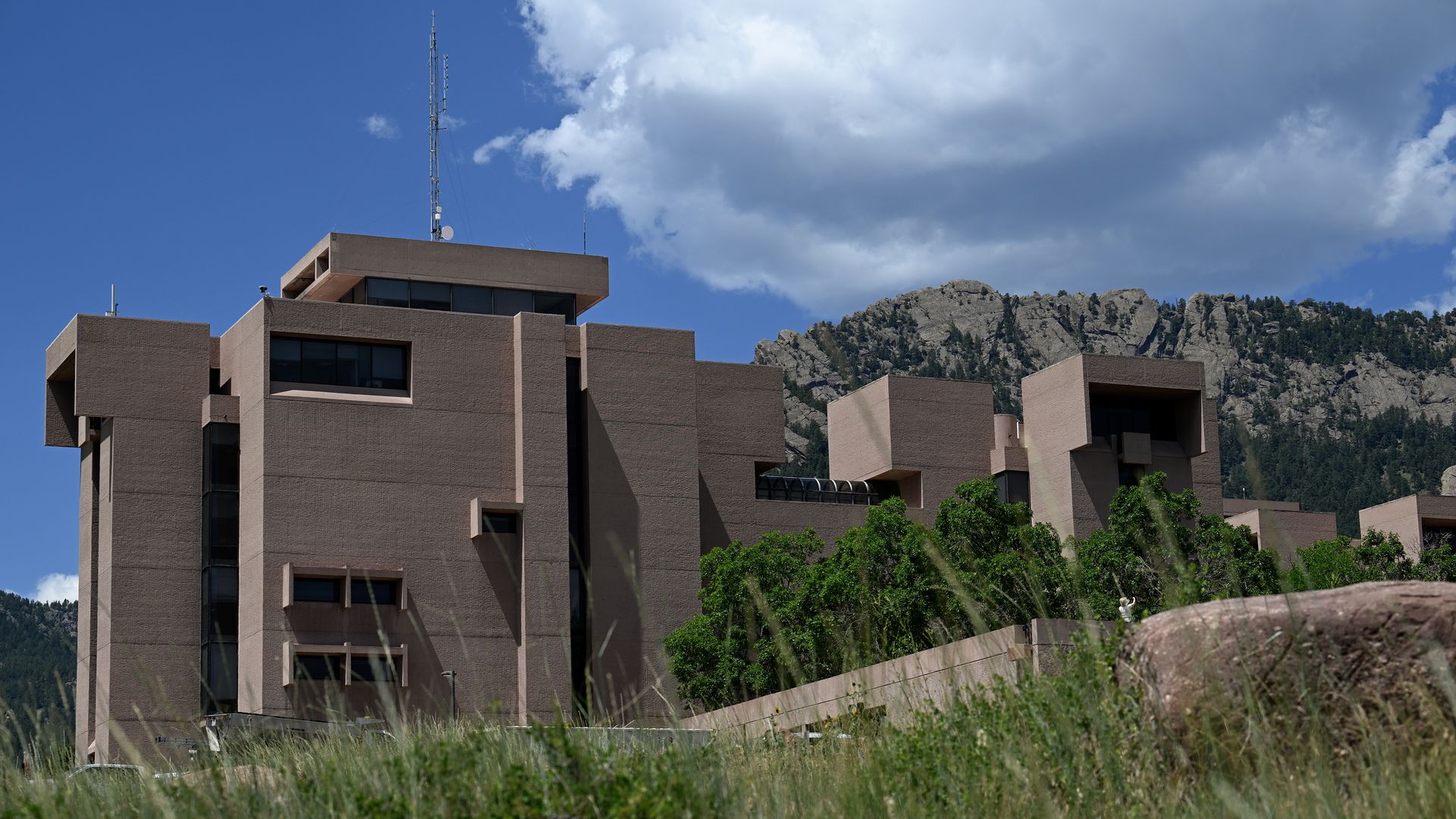 Large brown concrete building with many rectangular sections and windows, surrounded by green trees and grass, set against rocky mountains and a blue sky with white clouds.