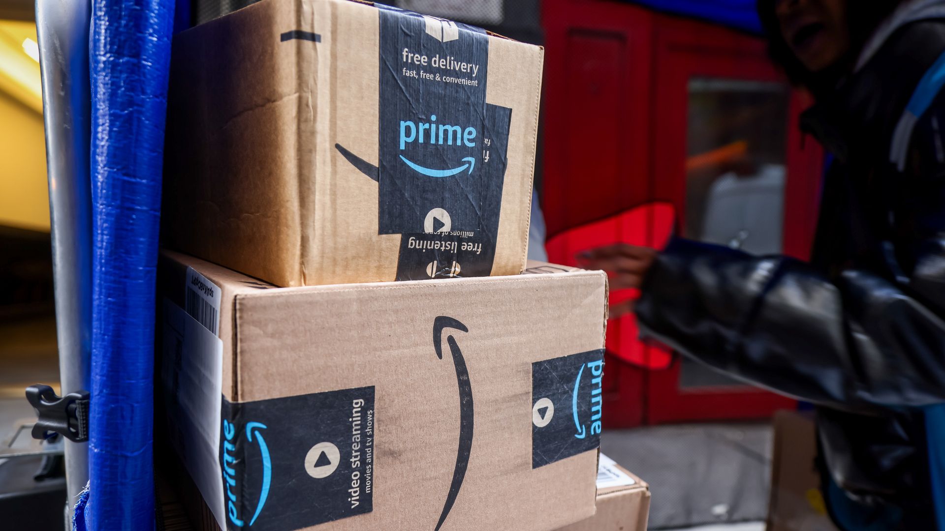 Stack of brown Amazon Prime delivery boxes secured with black tape bearing the blue Prime logo, inside a vehicle with a person in a black jacket in the background.