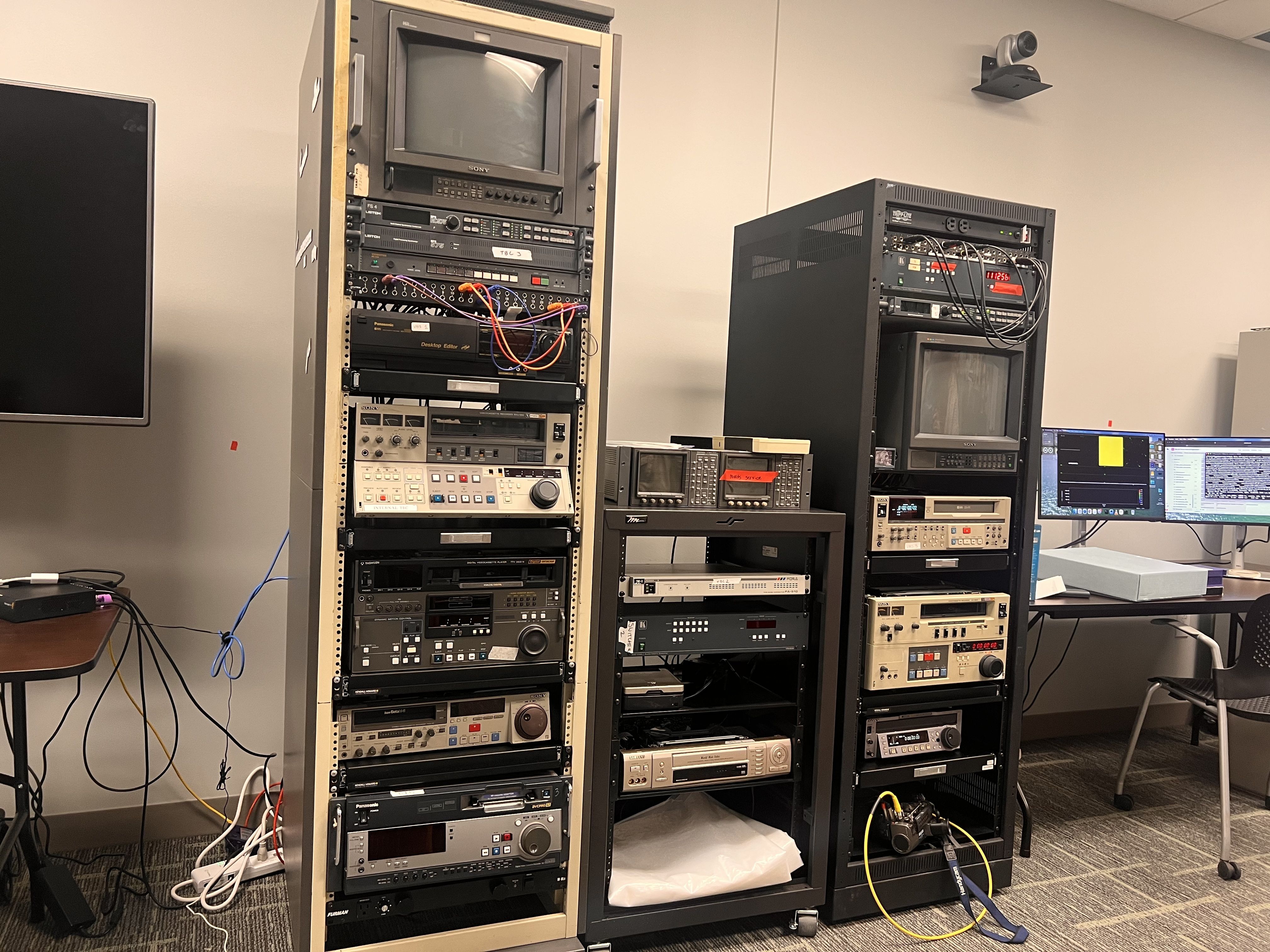 Two tall racks of video and audio editing equipment with numerous buttons, dials, and cables in an office room with monitors and a chair nearby.