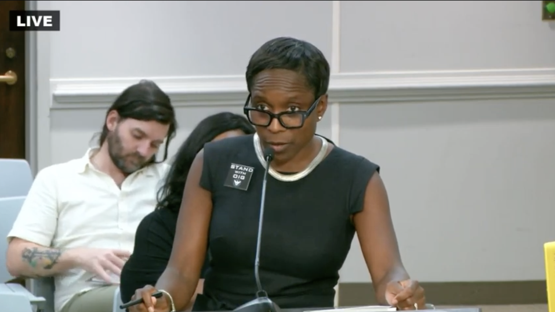 An individual wearing a button that says "STAND WITH OIG" speaks at a lectern during a government meeting