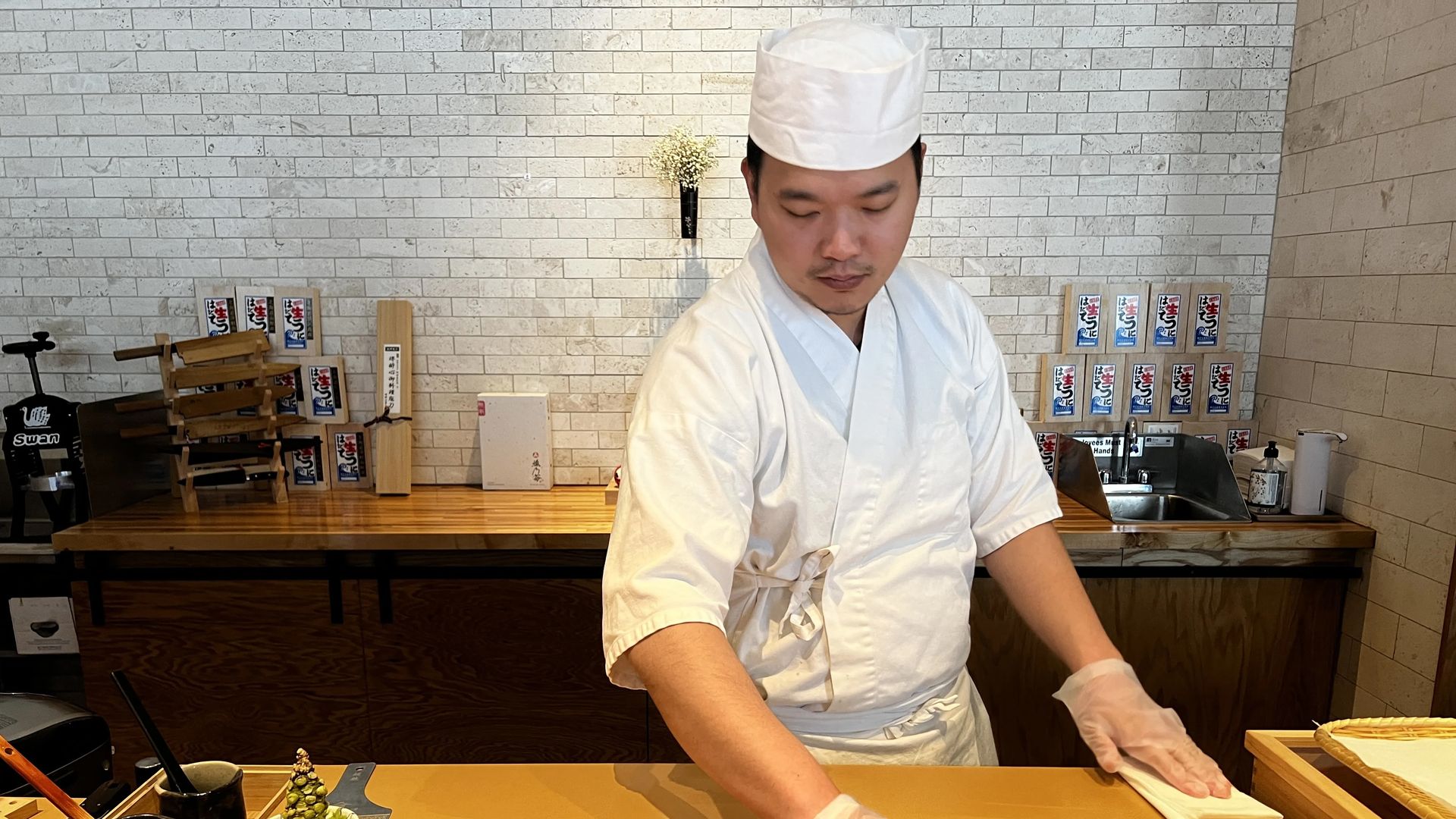 Sushi chef in white uniform and hat prepares dishes behind wooden counter with knife, small bowls of seafood, and condiments in a restaurant with light brick wall background.
