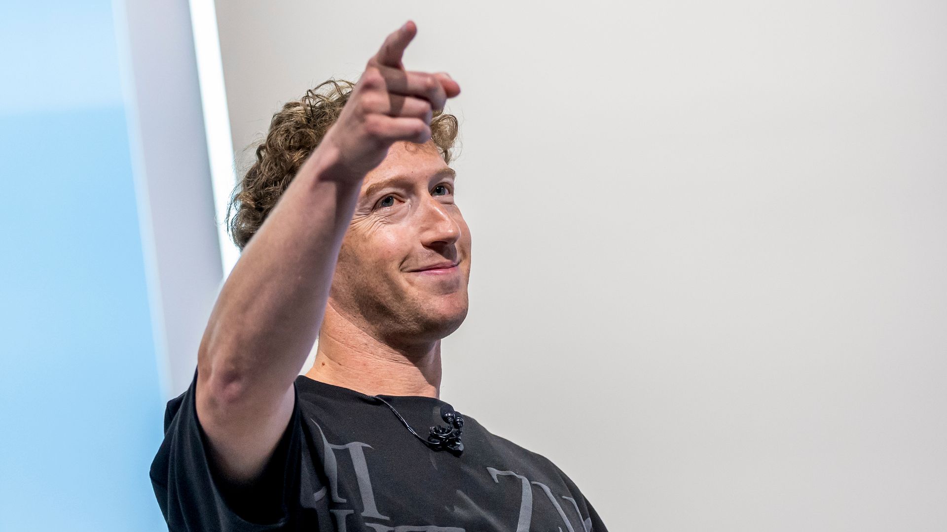 Presenter with curly hair in a black shirt raises his right hand and points upward, speaking on stage; blue panel at left and pale background.