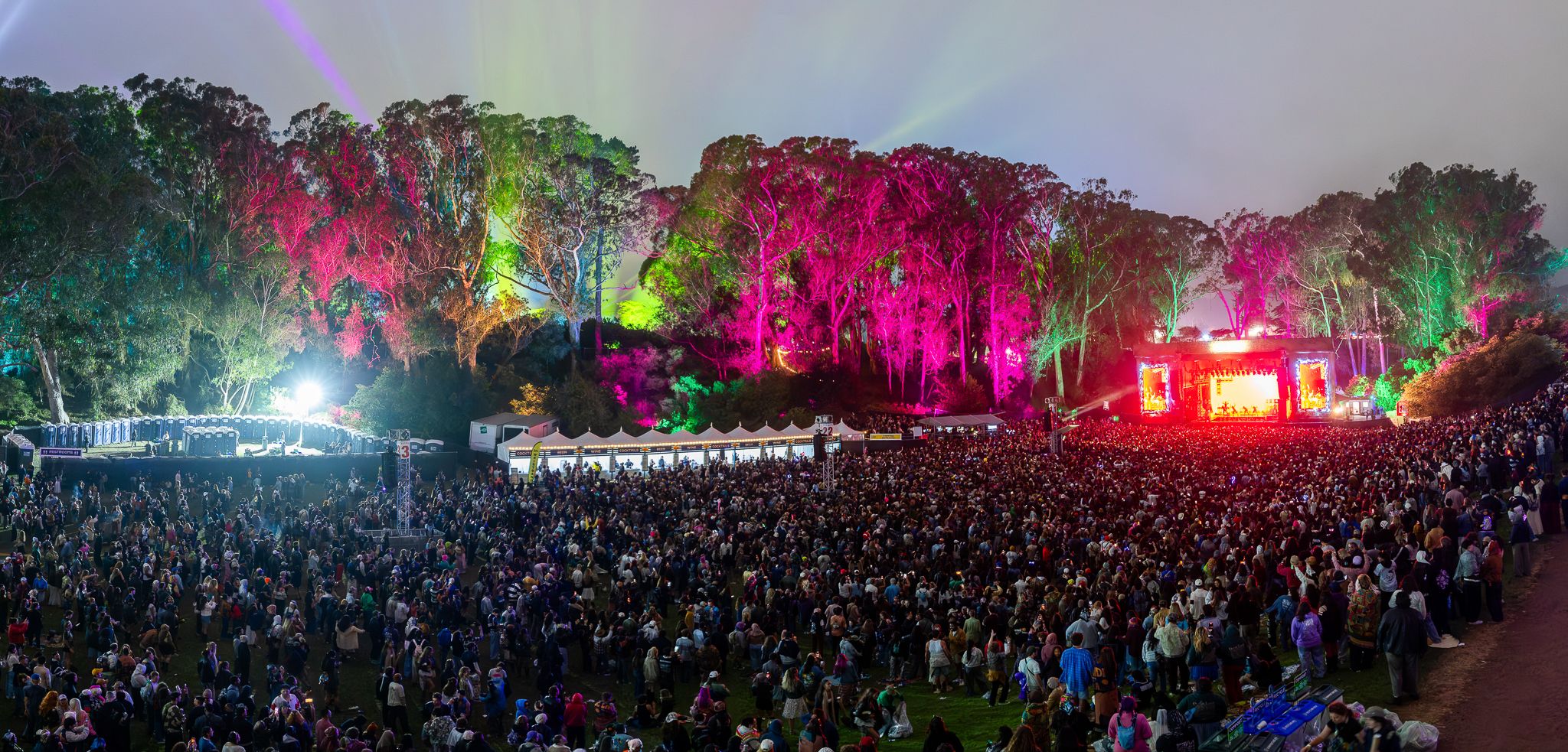 A large outdoor concert at night with a massive crowd facing a brightly lit stage on the right; colorful lights illuminate the trees and sky in pink, green, and purple.