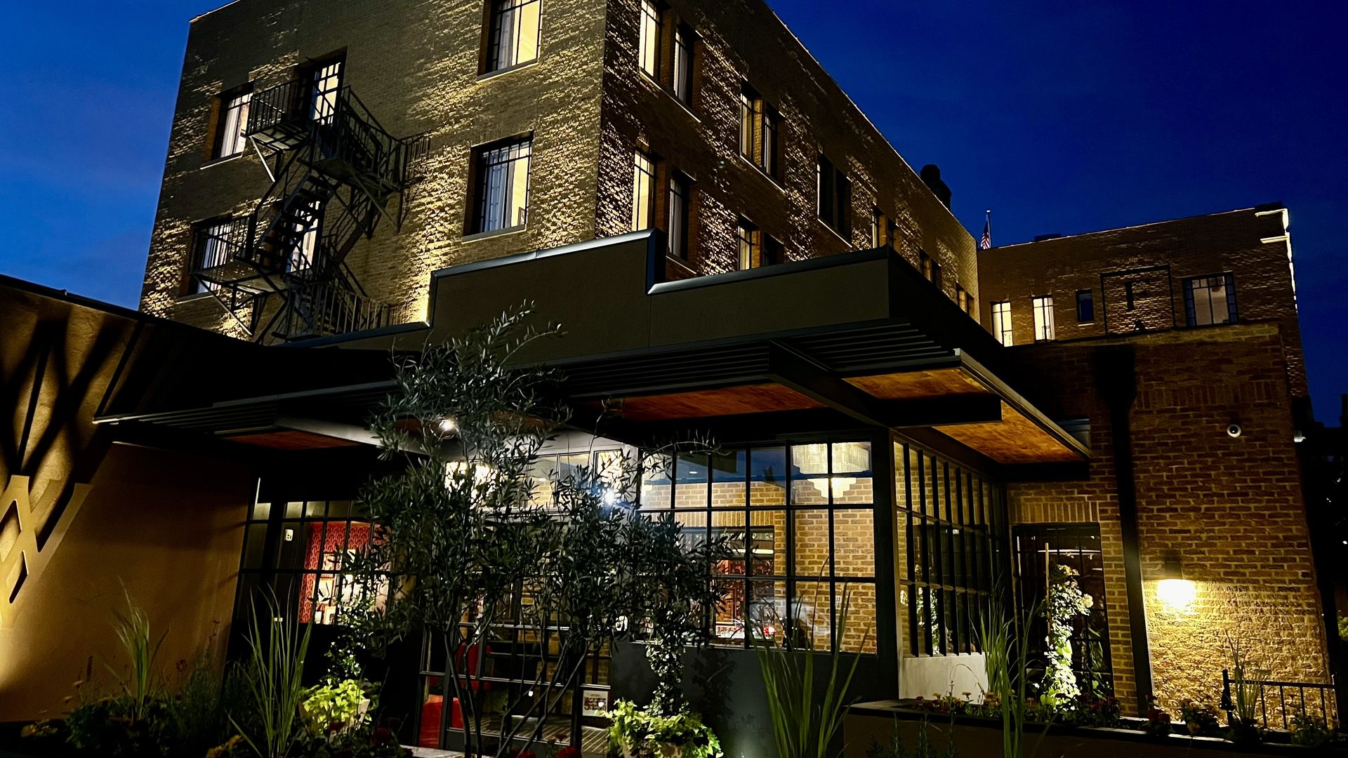 Nighttime exterior of a brick corner building with a metal fire escape on the left, warm-lit windows, a glass storefront at street level, and plants in the foreground against a deep blue sky.