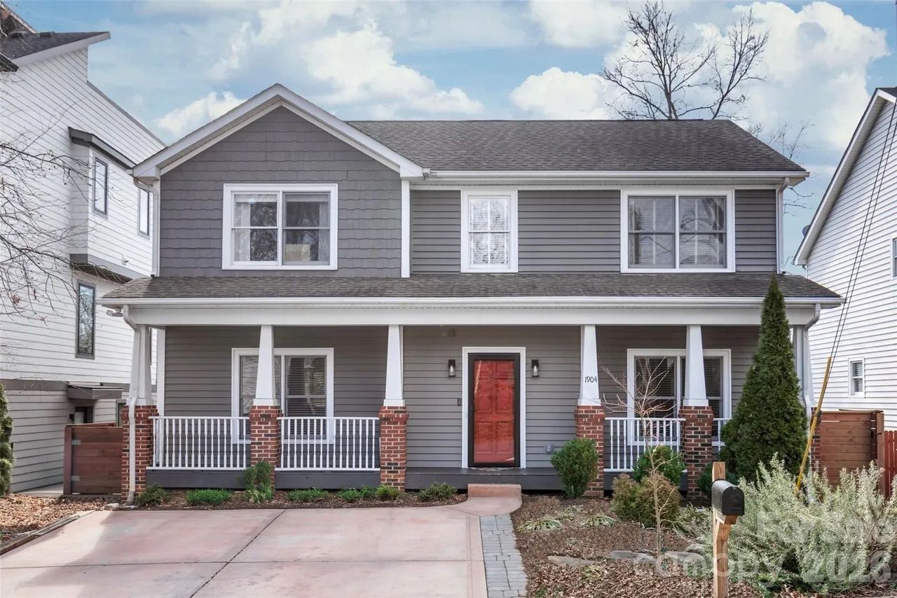 Two-story gray house with white trim, red front door, brick pillars on porch, driveway, and landscaped yard under a blue sky with clouds.