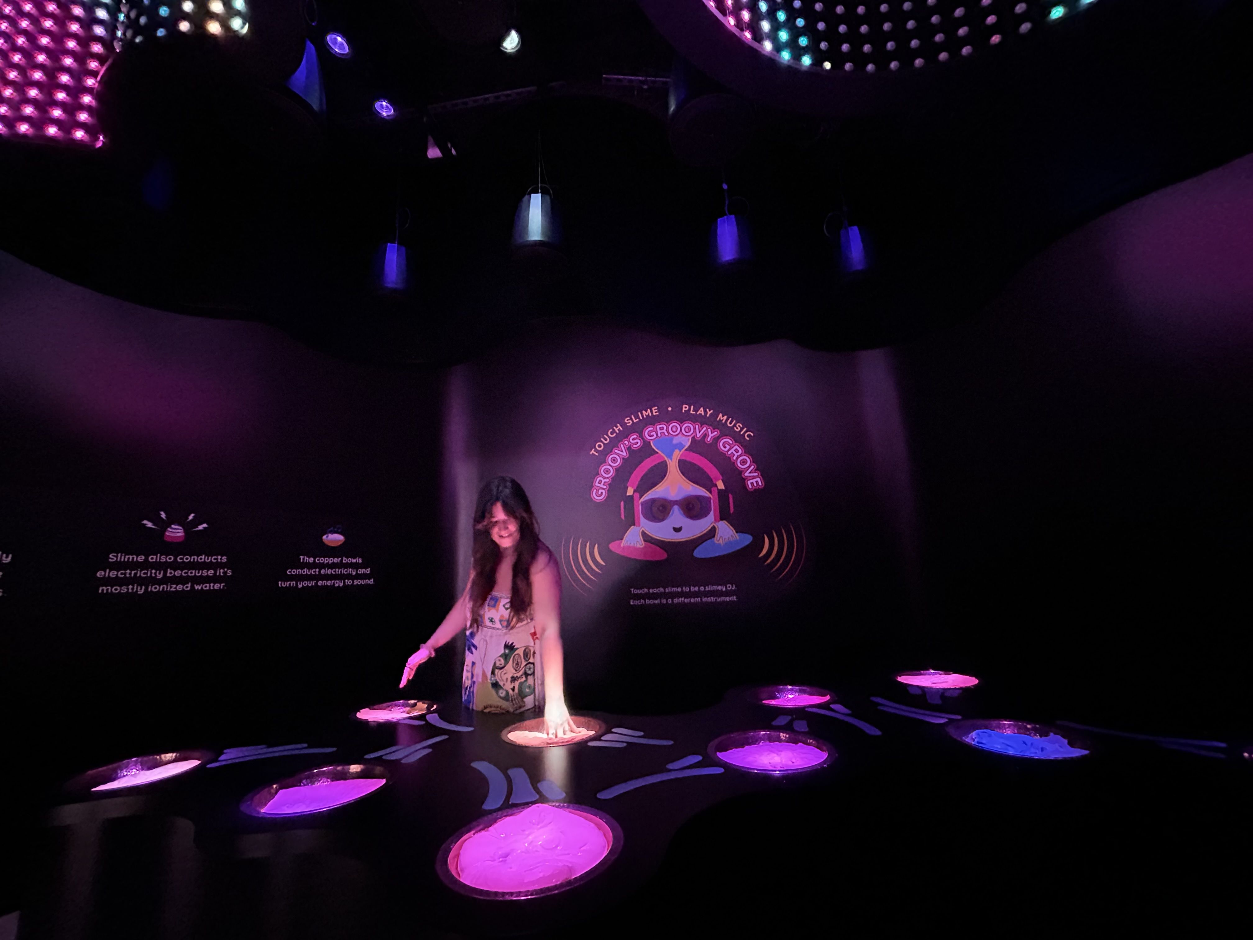 Dim exhibit space with purple lighting. A woman in a colorful dress stands on glowing pink floor circles; wall art shows a cartoon creature wearing headphones and the neon sign "Groovy Groove".