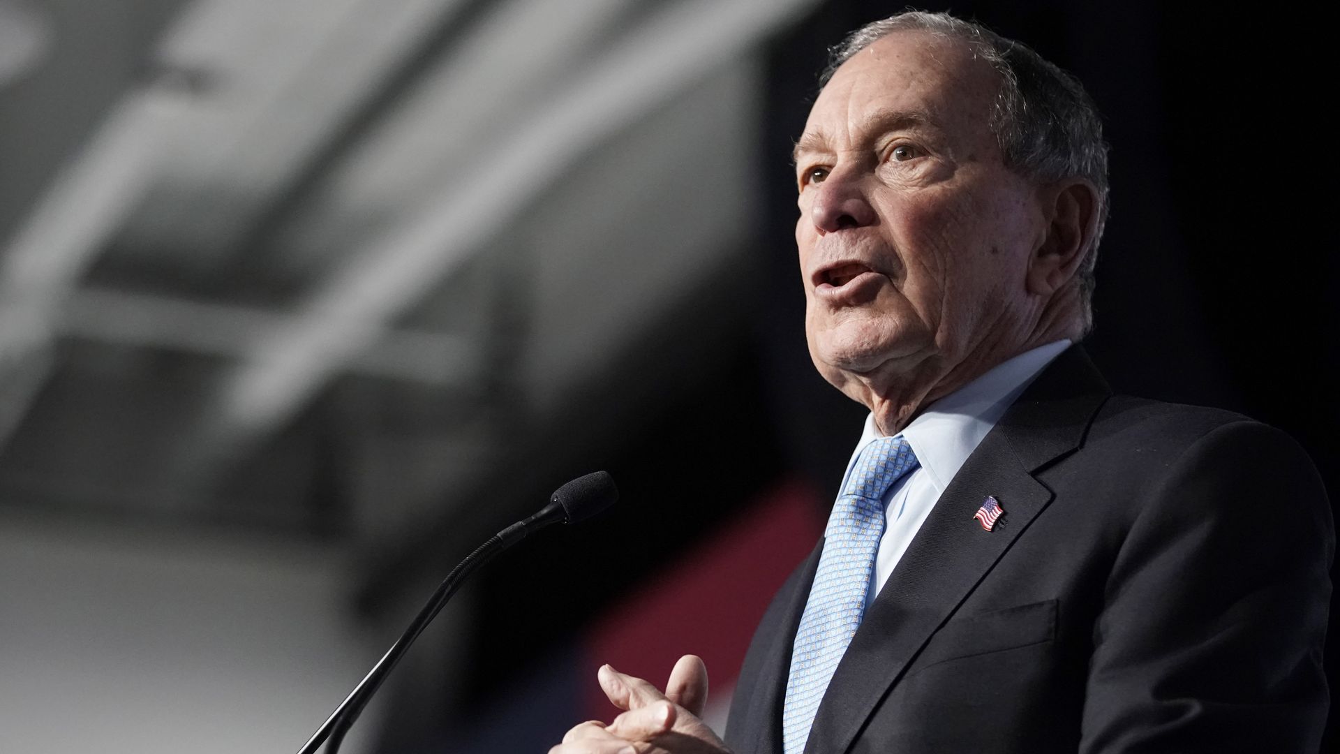 In this image, Bloomberg stands and talks in a suit