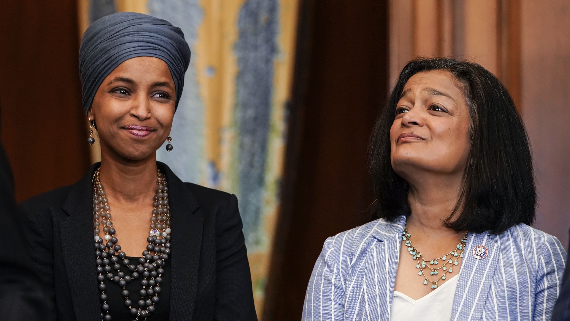 Rep. Ilhan Omar, wearing a blue headscarf, black blazer and black shirt with beaded necklaces, and Rep. Pramila Jayapal, wearing a baby blue blazer with white stripes, a white shirt and a turquoise necklace.
