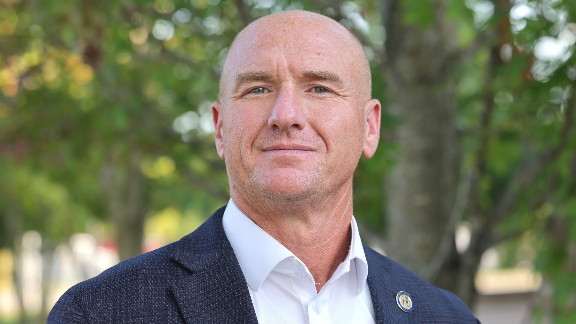 A bald man in a navy plaid suit jacket and white dress shirt stands outside in front of green, leafy trees, smiling slightly.