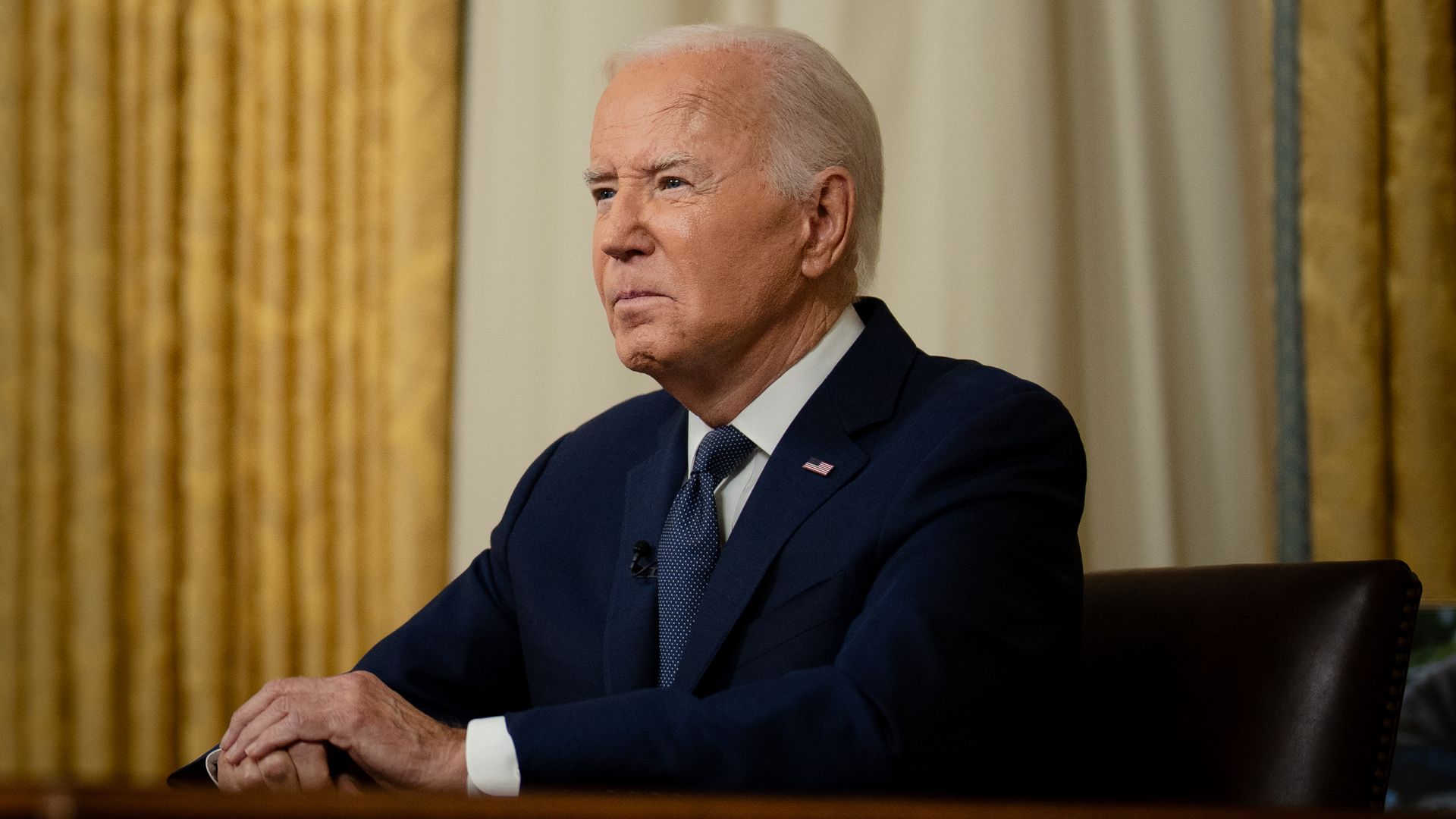 Joe Biden, wearing a blue suit and sitting at the resolute desk in front of gold curtains.