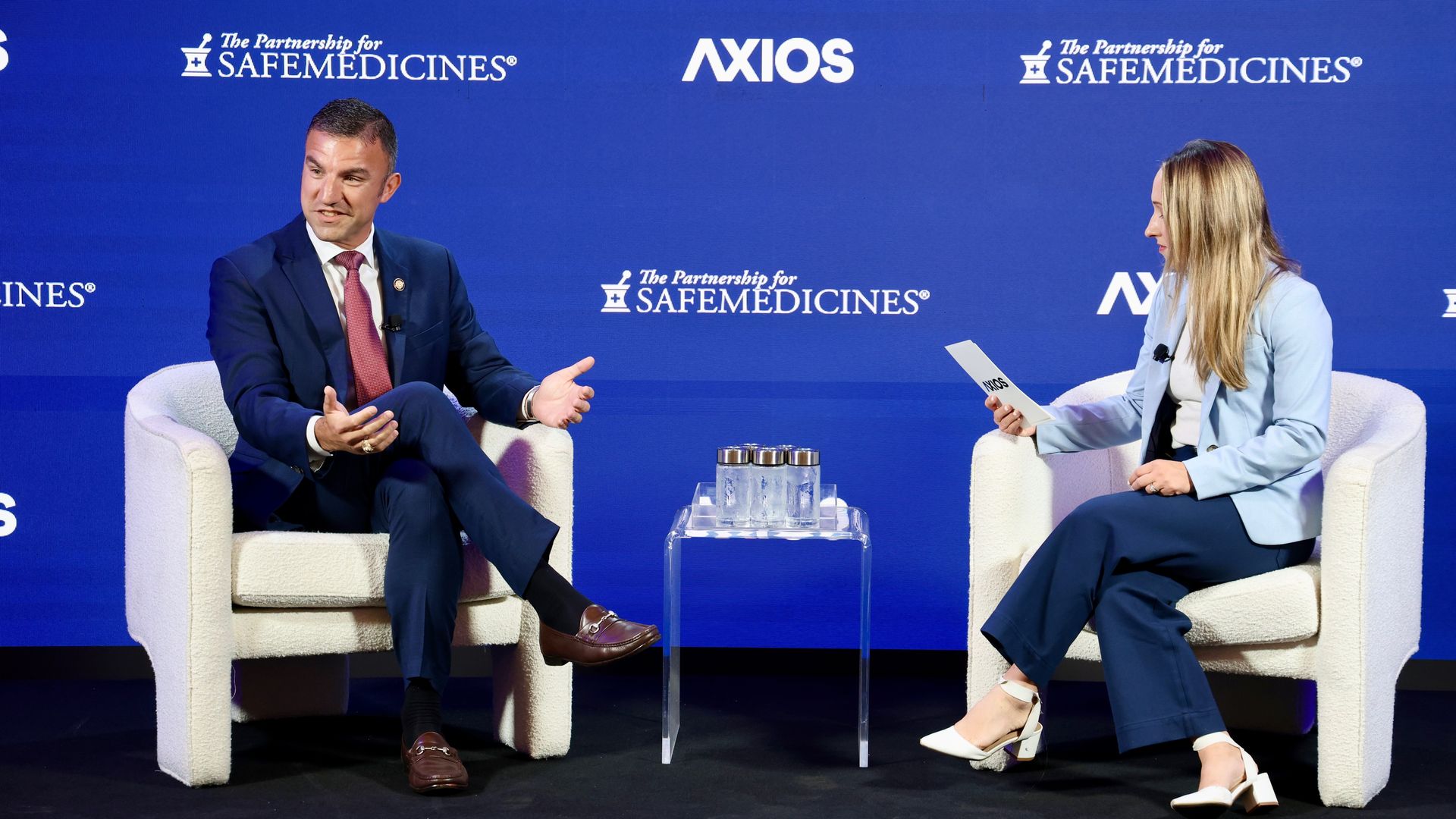 Two panelists sit on blue stage with logos; Rep. Rudy Yakym (R-Ind.) in a dark suit and red tie on the left and Axios congressional reporter Stef Kight in a light blue blazer on the right, with a glass table and water between them, Axios backdrop.
