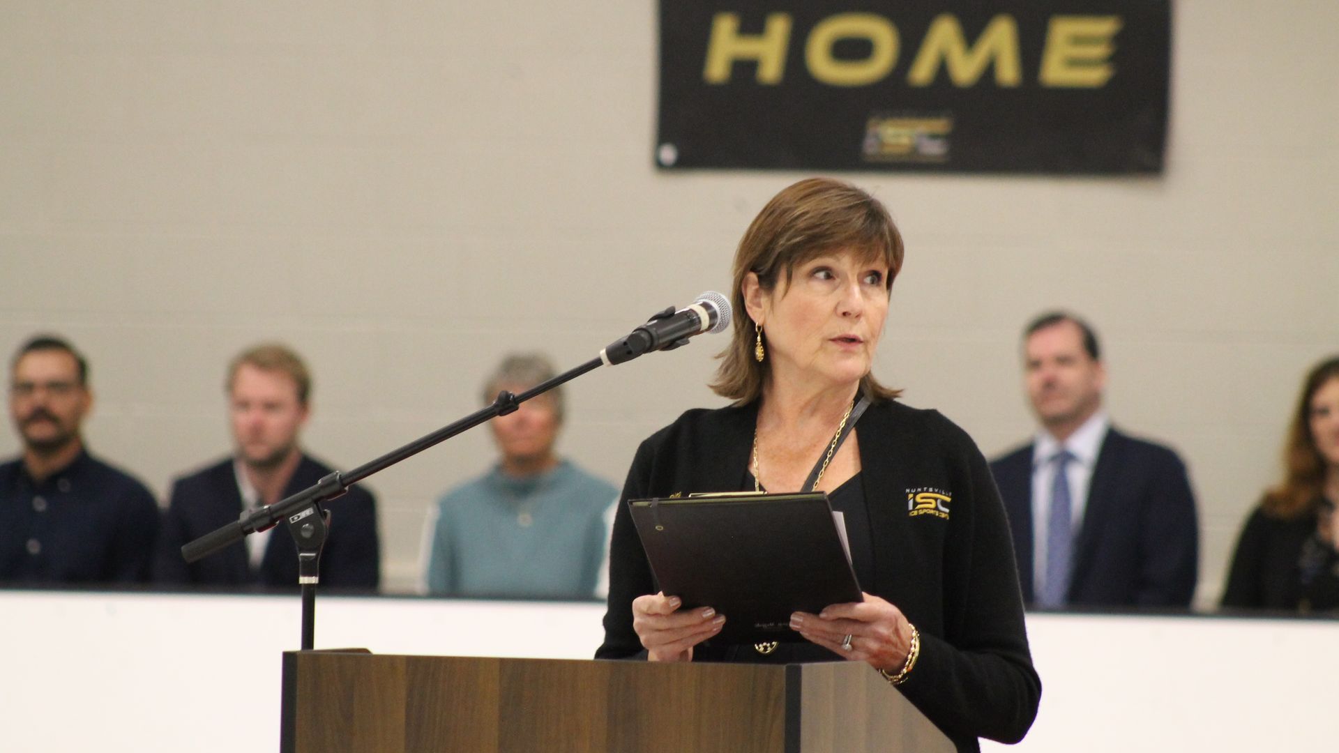 A woman in black with a clipboard speaks at a podium with microphone in a gym or event space, with a black sign reading HOME in yellow letters behind her and people seated in the background.