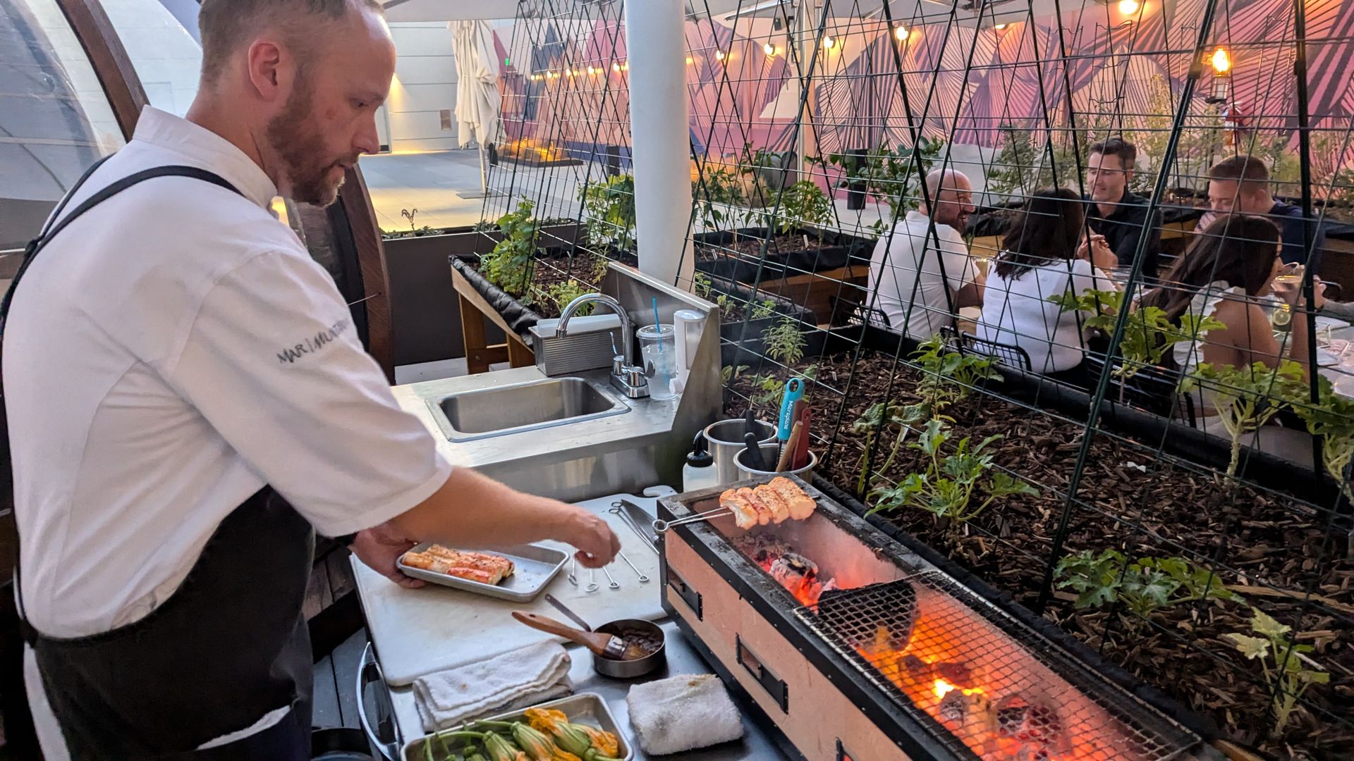 A cook grills a skewer of crab legs over hot coals on a rooftop patio.