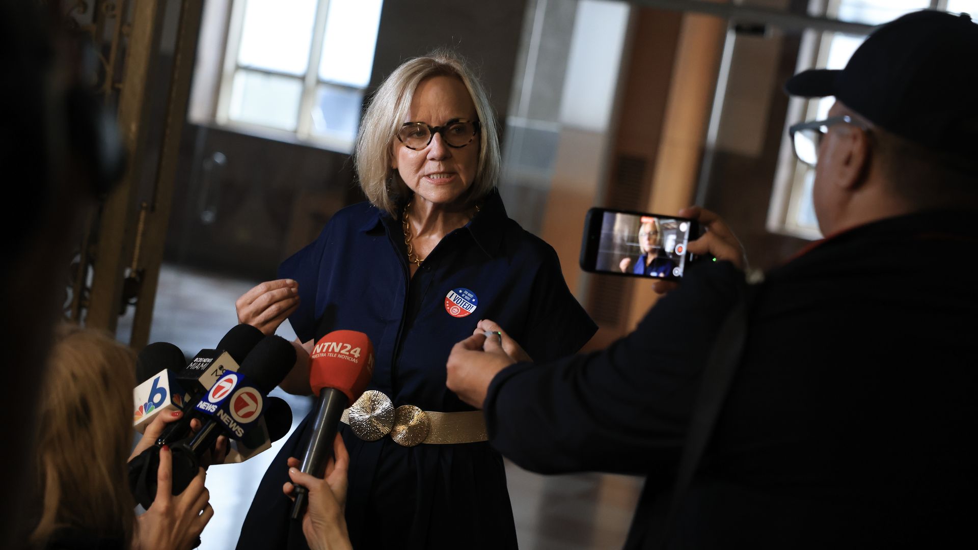 A blonde haired woman in glasses wearing a dark top speaks to reporters standing in front of her holding microphones and cameras.