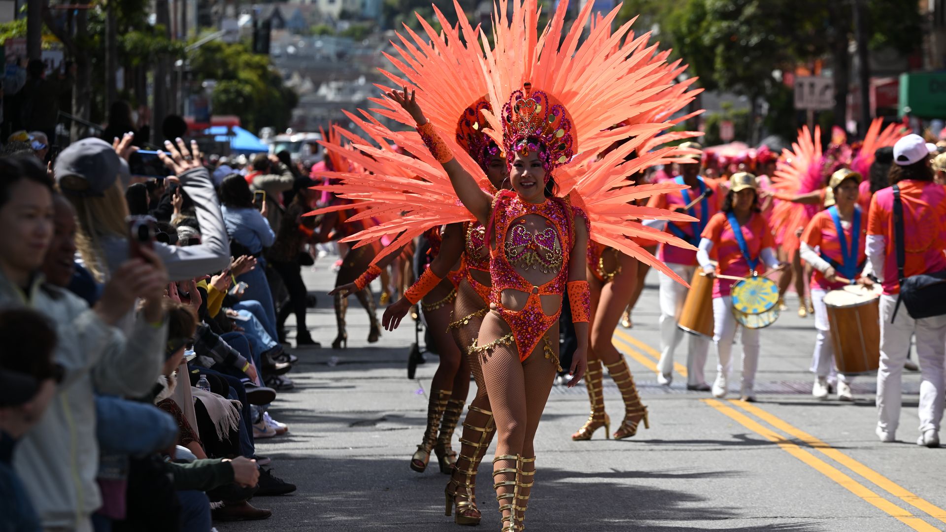Parade on city street with a central dancer in bright orange-pink costume and large feather headdress, smiling, flanked by drummers. Crowded audience on both sides captures the moment.