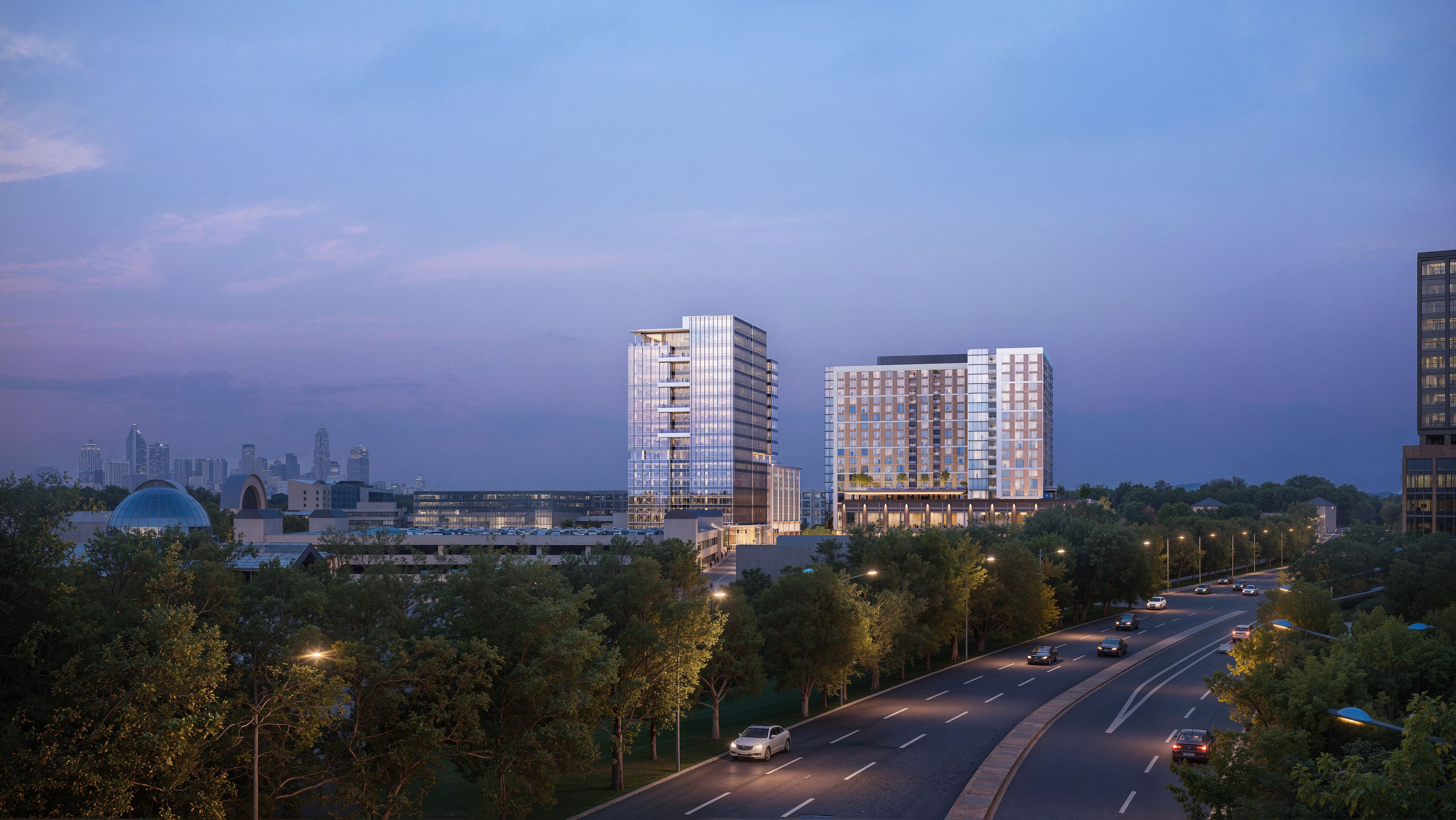 City skyline at dusk with glass office towers along a wide road, passing cars, and trees in the foreground; a blue-domed building sits on the left under a blue-purple sky.