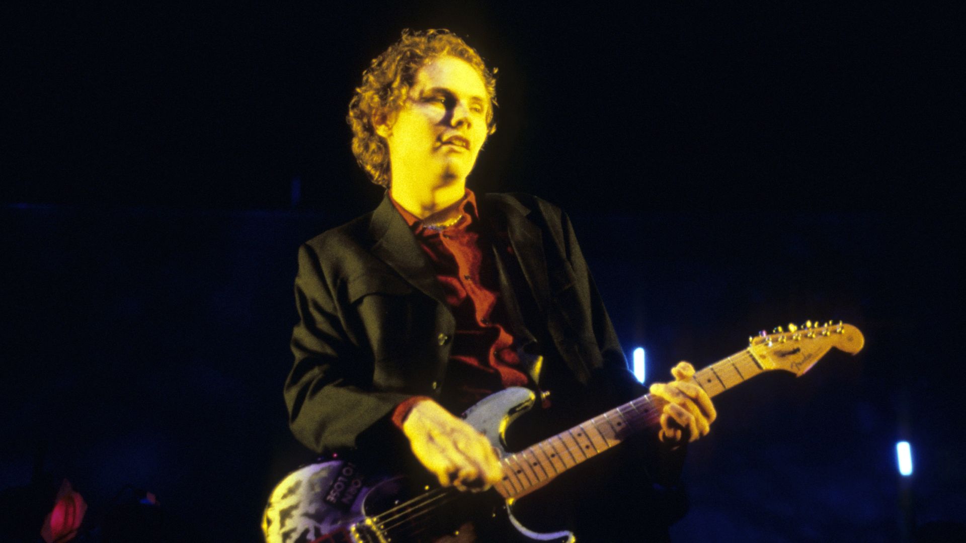 Guitarist on a dark stage, bathed in yellow light, wearing a black blazer over a red shirt, playing a light-colored electric guitar adorned with stickers.
