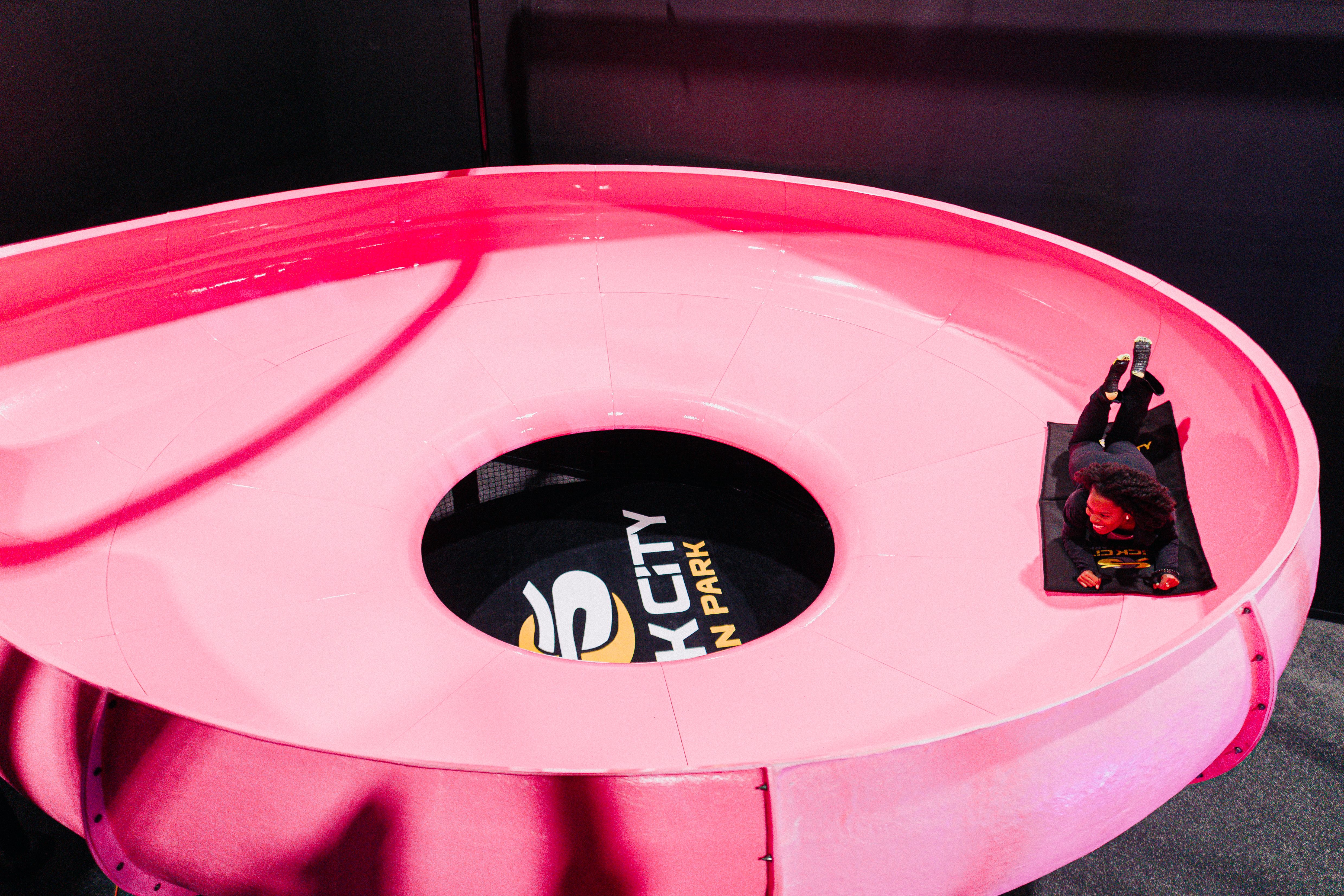 Donut-shaped bright pink amusement ride with a black center and white lettering; a person in dark clothing sits on the rim at the right, holding a small device.