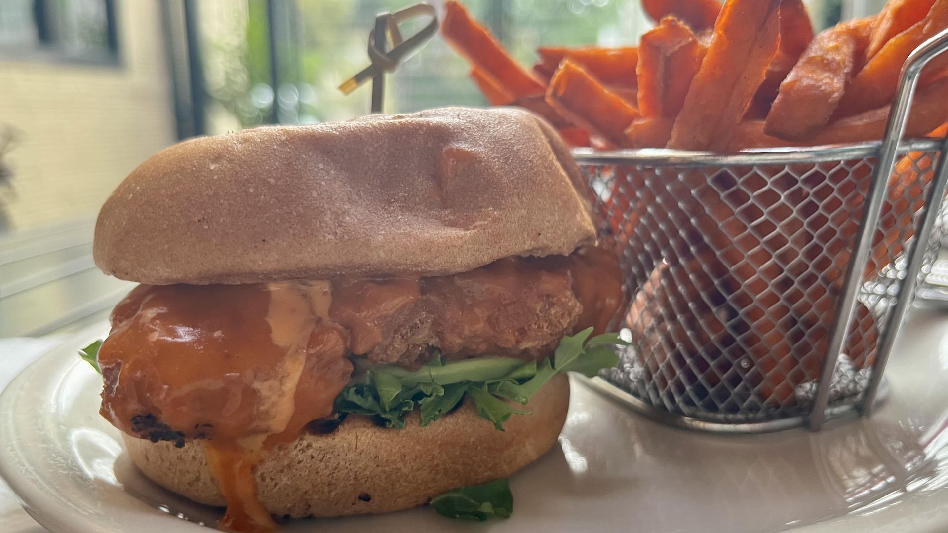 A vegan chicken burger on a plate overflows with buffalo sauce. Sweet potato fries are seen in the background.