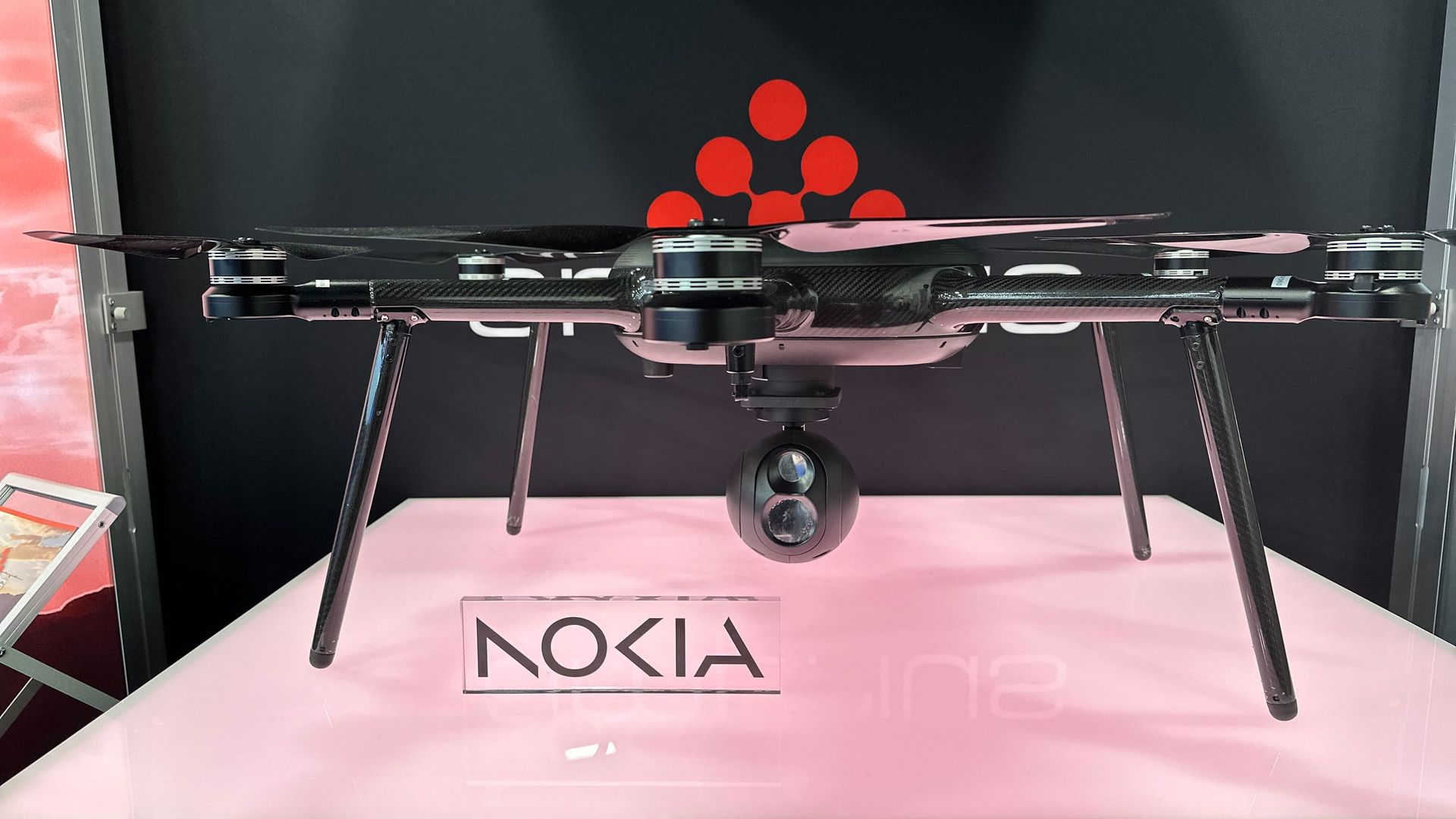 Black quadcopter drone with four propellers and a dual-lens camera mounted on a pink surface, displayed in front of a black backdrop with red dots and partially visible "Nokia" sign.