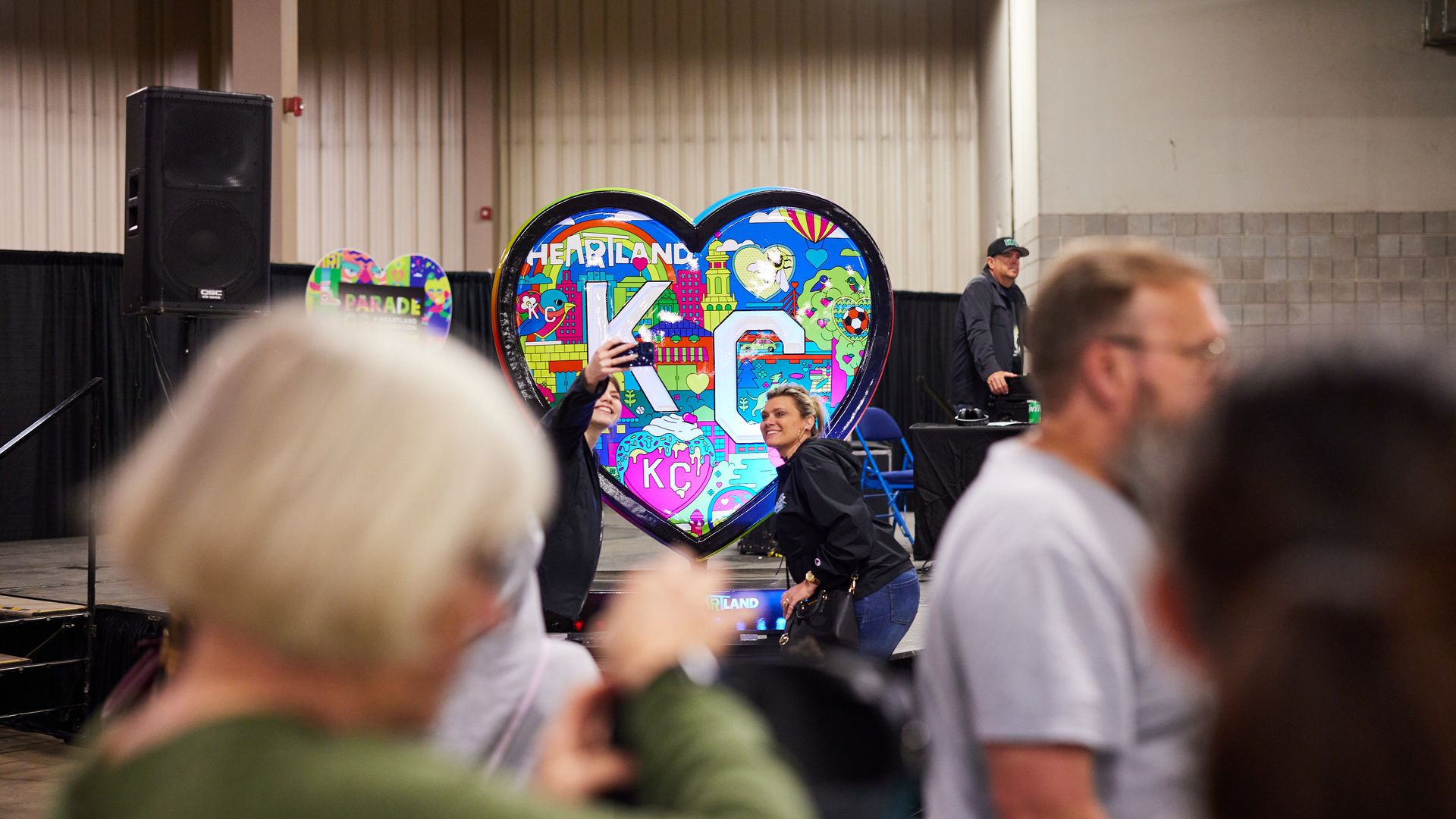 Two people stand in front of a colorful heart that says KC in the middle.