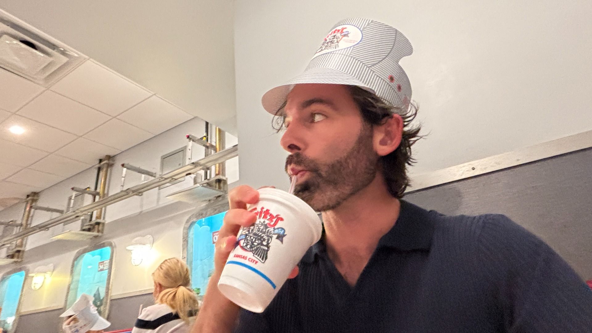 Bearded man in a light, striped hat sips from a large white cup with a straw in a retro diner, with blue-walled display cases and metal shelving in the background.