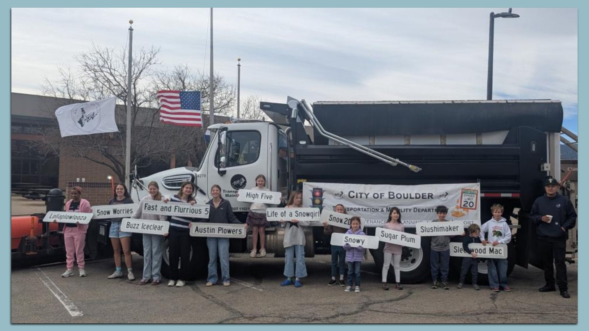 Group of people, mostly children, stand in front of a black and white City of Boulder truck holding signs with winter-themed names like "Snowy" and "Sugar Plum," with American and white flags flying.