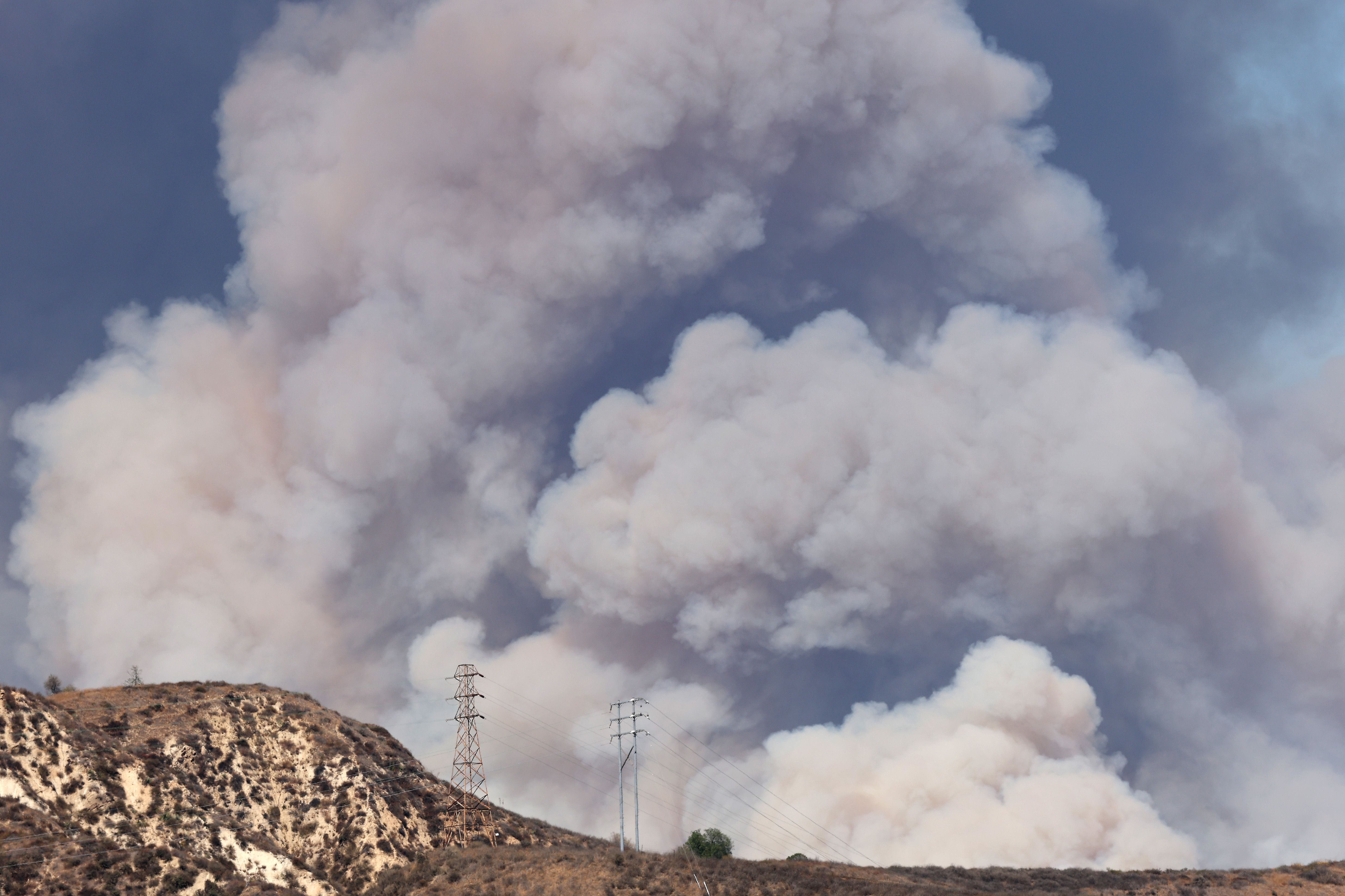 Large clouds of thick smoke rising from a wildfire burning in dry hills with sparse vegetation and two electrical transmission towers visible.