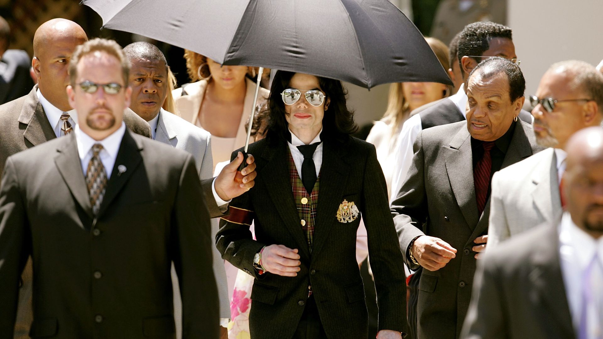 A crowd of formally dressed people walking outdoors; at center, Michael Jackson in a black suit, plaid vest and tie wears sunglasses as someone holds a large black umbrella above her.