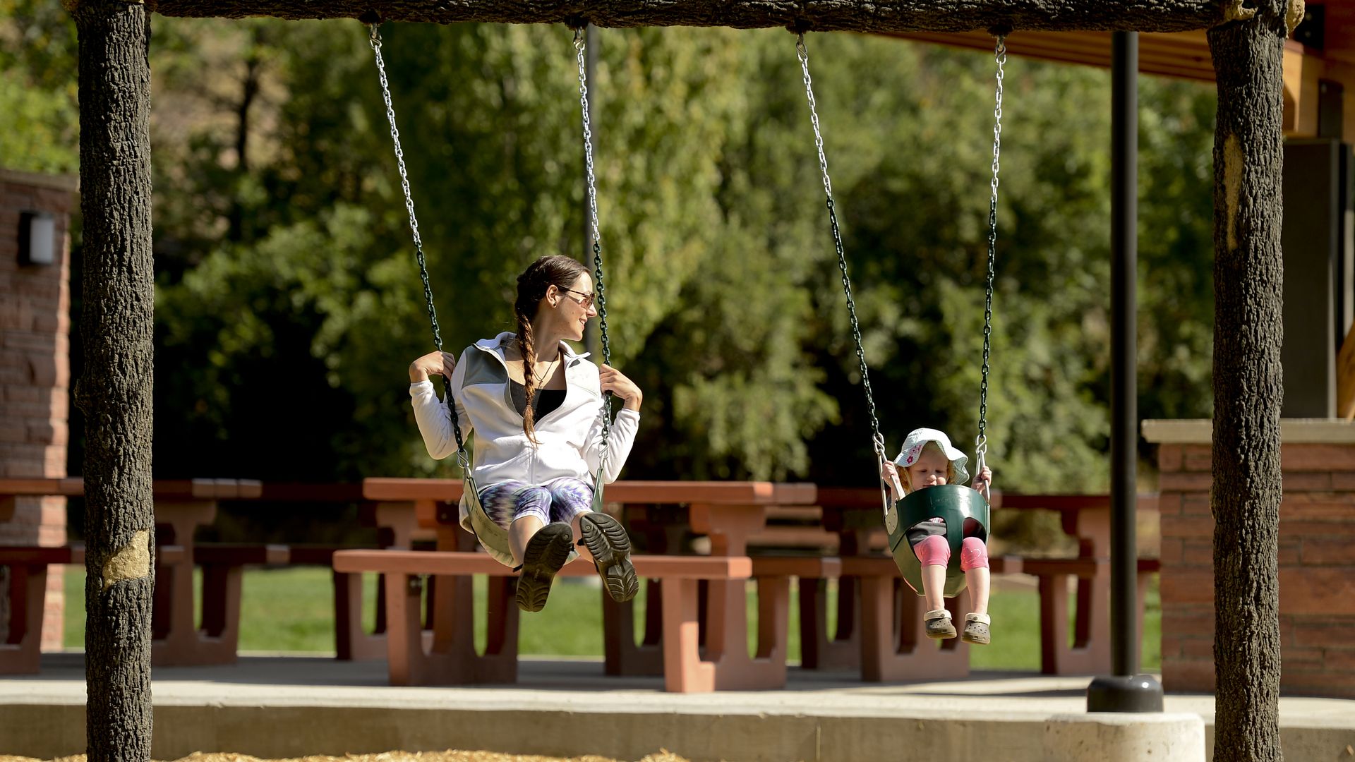 A woman and a toddler swing side by side on a swing set in a park with wood chip ground, picnic tables, and greenery in the background on a sunny day.
