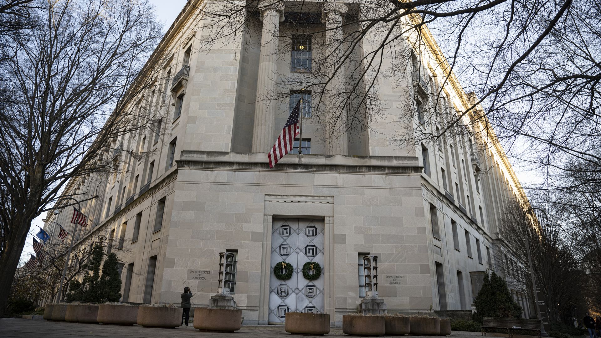 Stone government building with large columns and two wreaths on the door, an American flag, leafless trees in front under a clear sky in late afternoon light.
