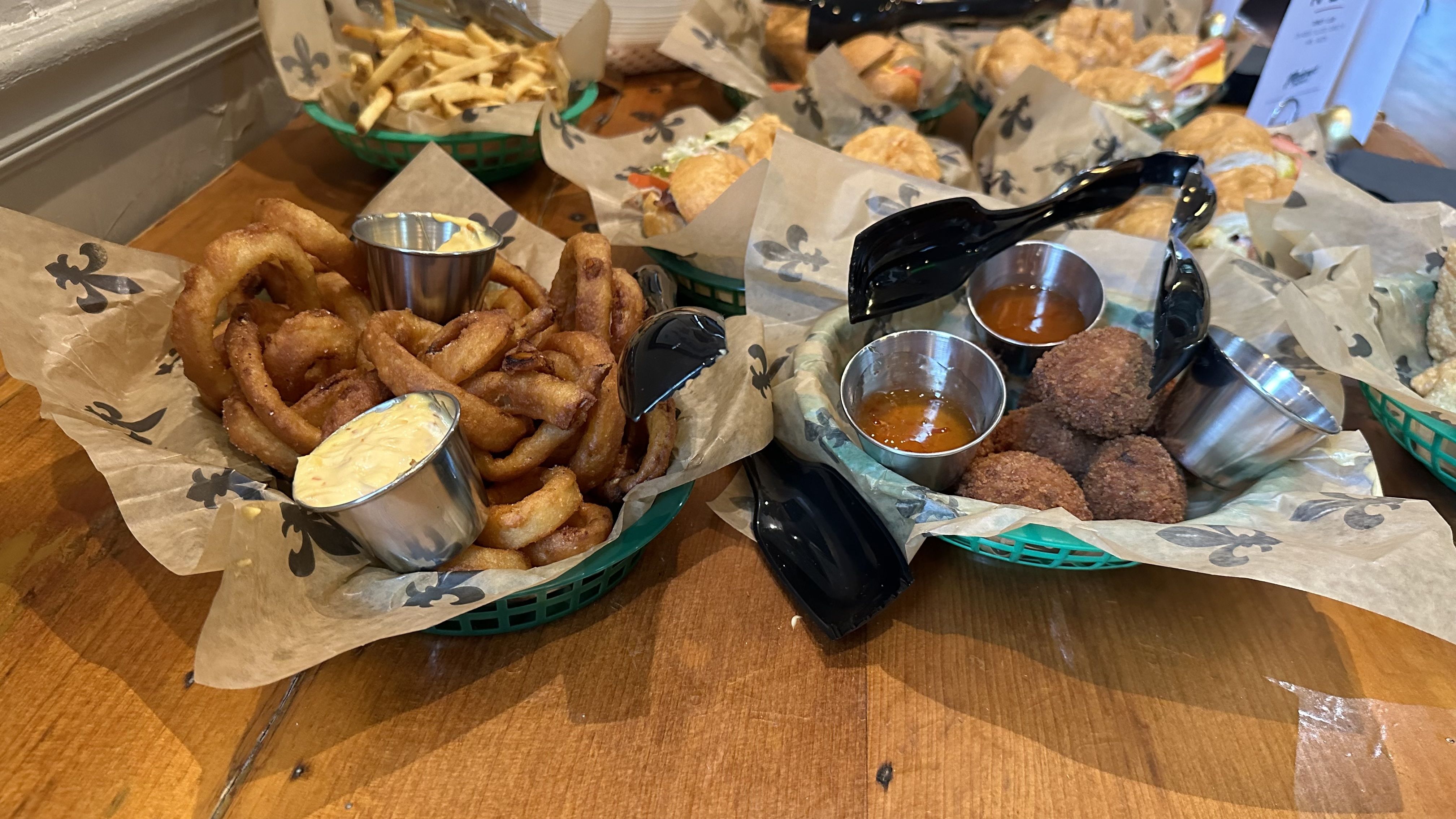 A basket of onion rings and a basket of boudin balls sit on a table with black plastic tongs sitting on top.