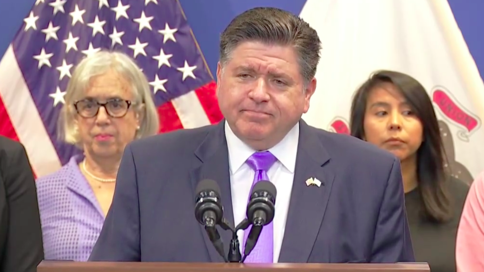Man in a blue suit and purple tie speaking at a podium with two people behind him and American and state flags in the background.