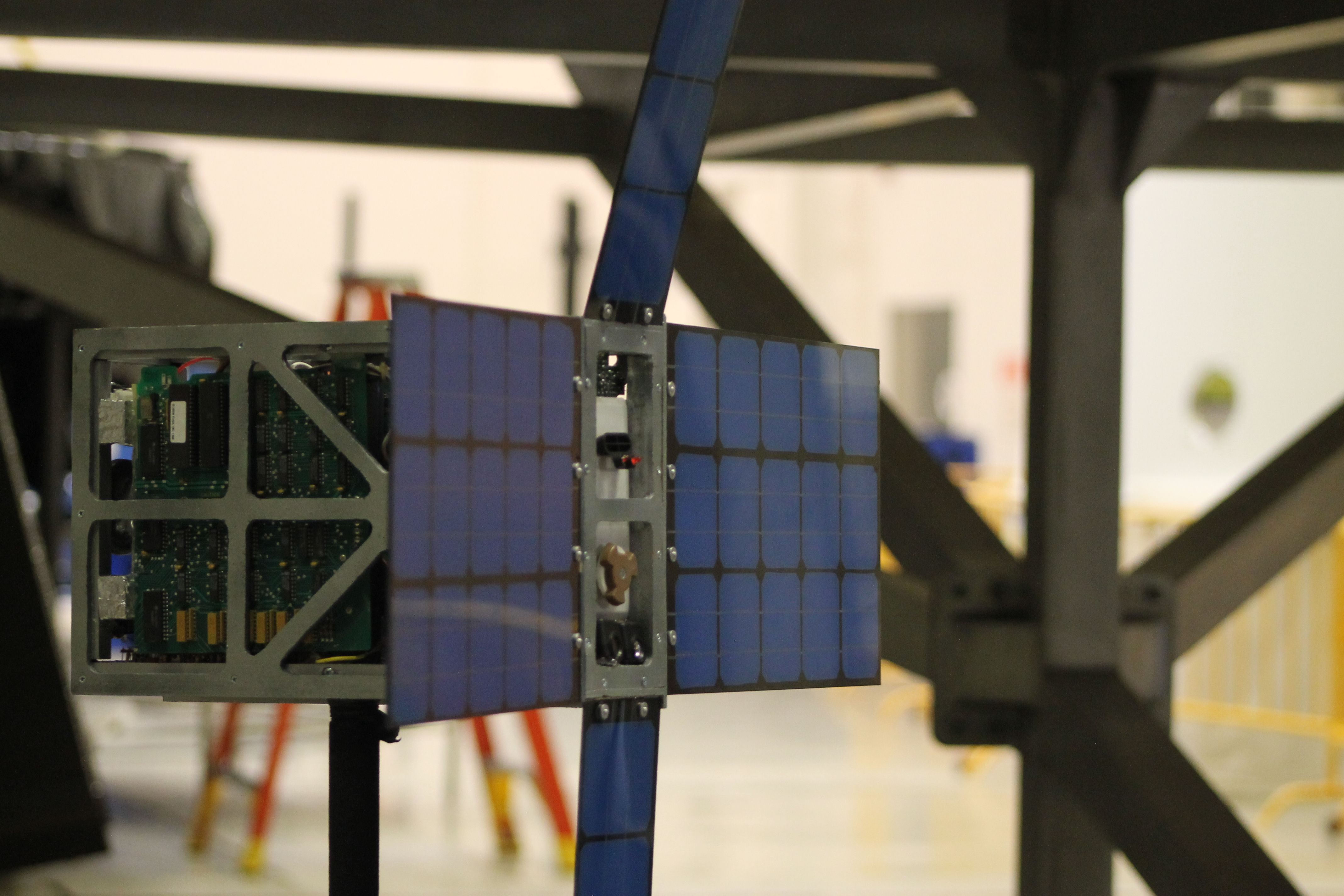 Close-up of a small satellite with blue solar panels and exposed internal components, set against an industrial workshop background with yellow ladders and metal framework.