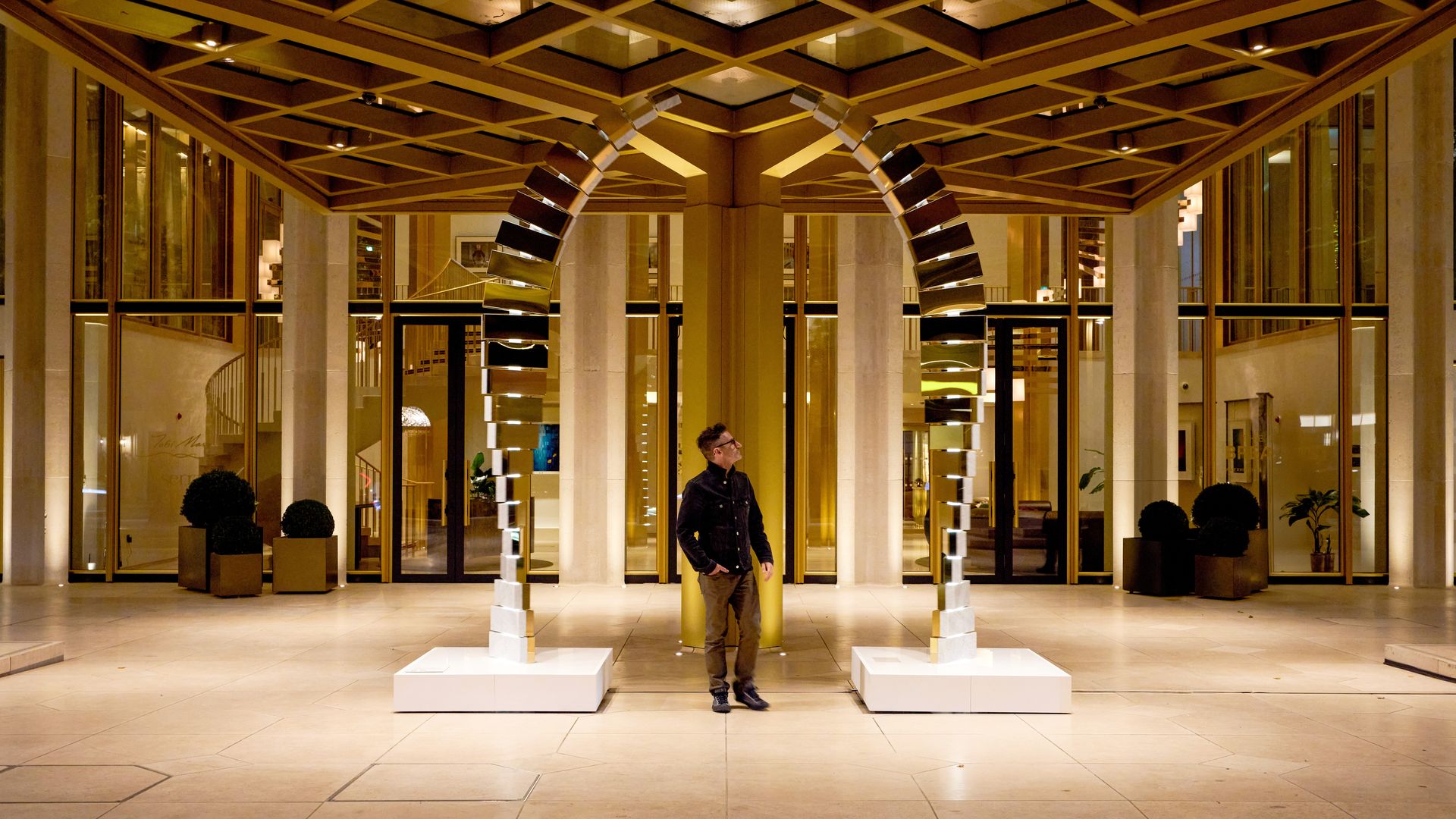 A man in dark clothes stands between two modern mirrored arch sculptures inside a spacious, golden-lit building with geometric ceiling patterns and large glass doors.