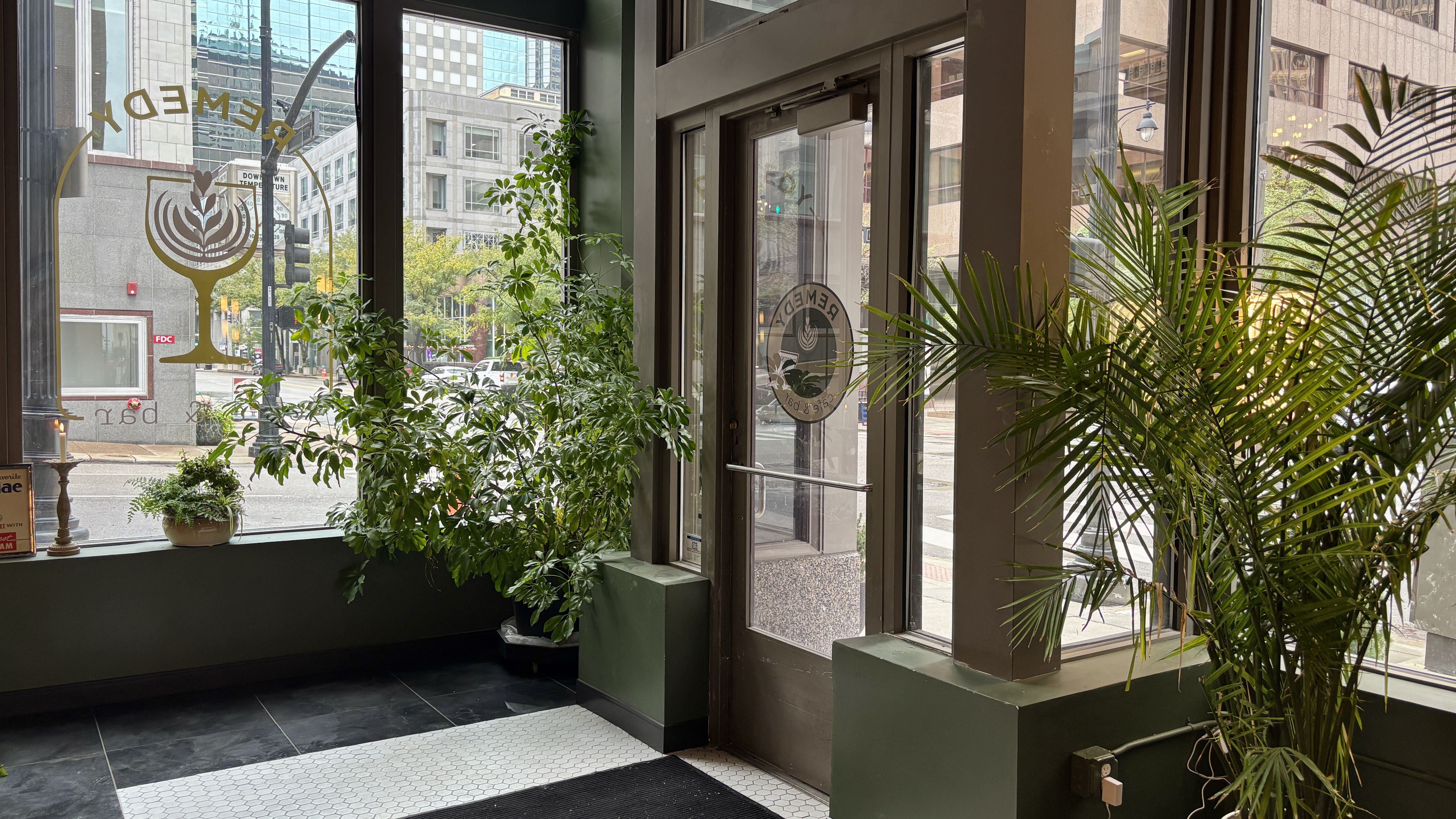 Indoor lobby with lush green plants in pots along a wide window. Black-and-white tiled floor, glass doors with a circular emblem, and a city street visible outside with buildings and a traffic light.