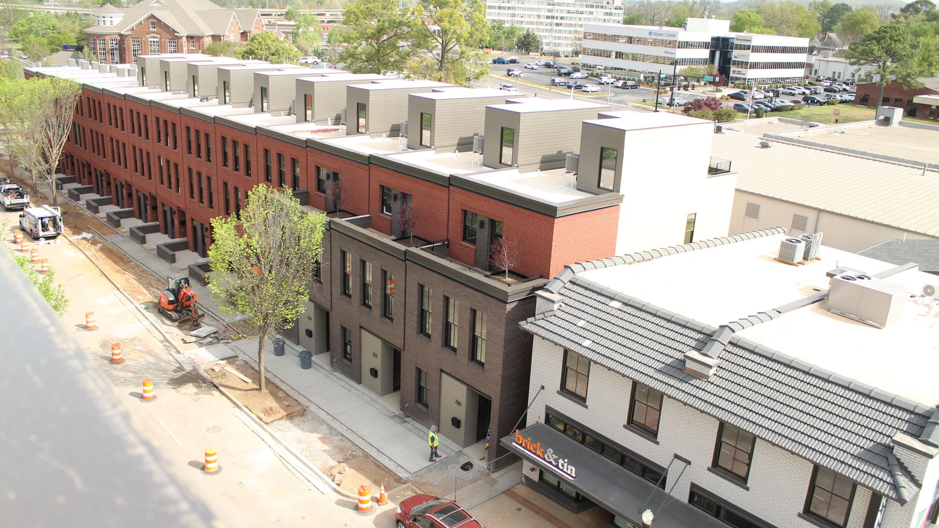 Aerial view of a street of red brick townhouses with gray rooftop dormers under construction. Orange cones, a small excavator, trees, and a white brick building with a storefront reading brick & tin.