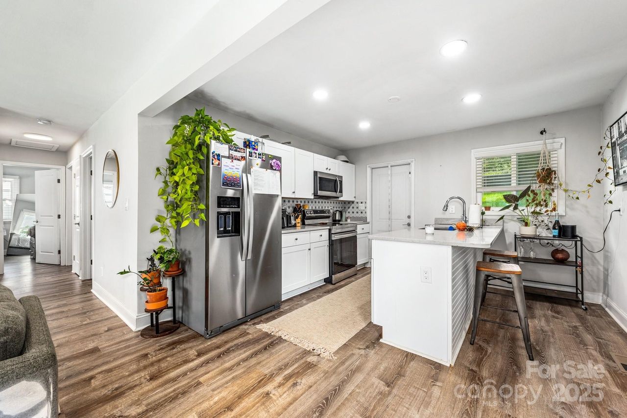Bright modern kitchen with white cabinets, stainless steel appliances, a large island with stools, wood flooring, and various green potted plants adding color and life.