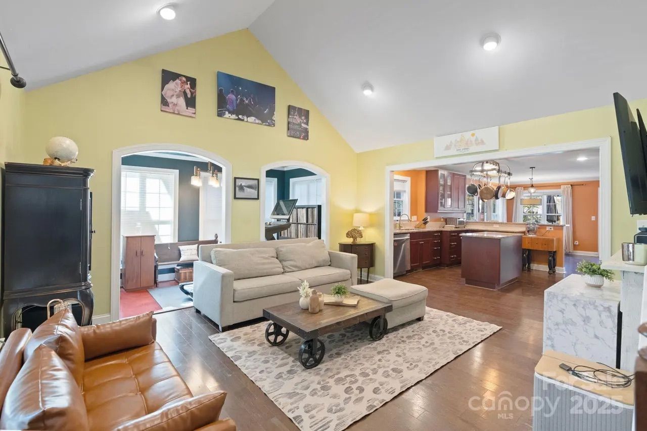 Living room with brown leather chair, light gray sofa, wooden coffee table on patterned rug, yellow walls, open view into kitchen with dark wood cabinets and orange walls, hanging pots.