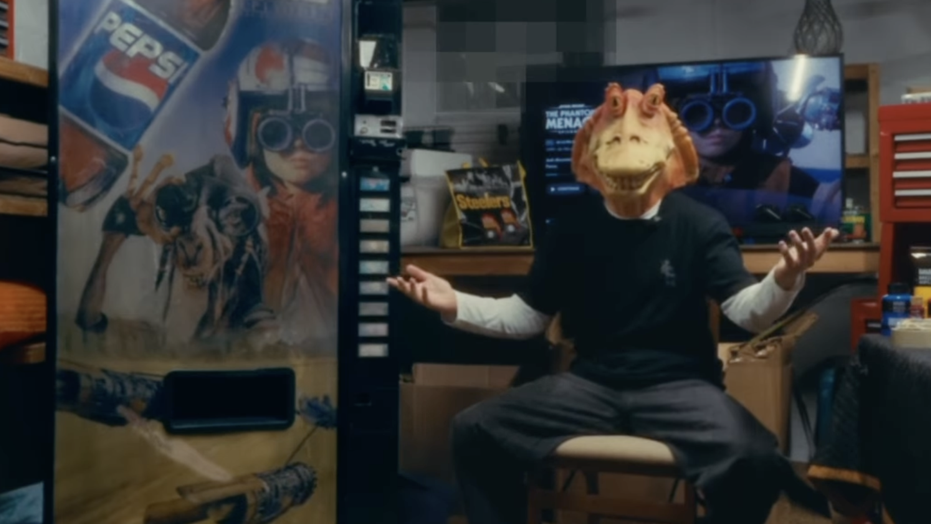 Person wearing a brown dinosaur mask sits on a stool in a cluttered workshop, arms raised. Left: large Star Wars/Pepsi poster; back: TV displaying a VR image; right: red tool chest.