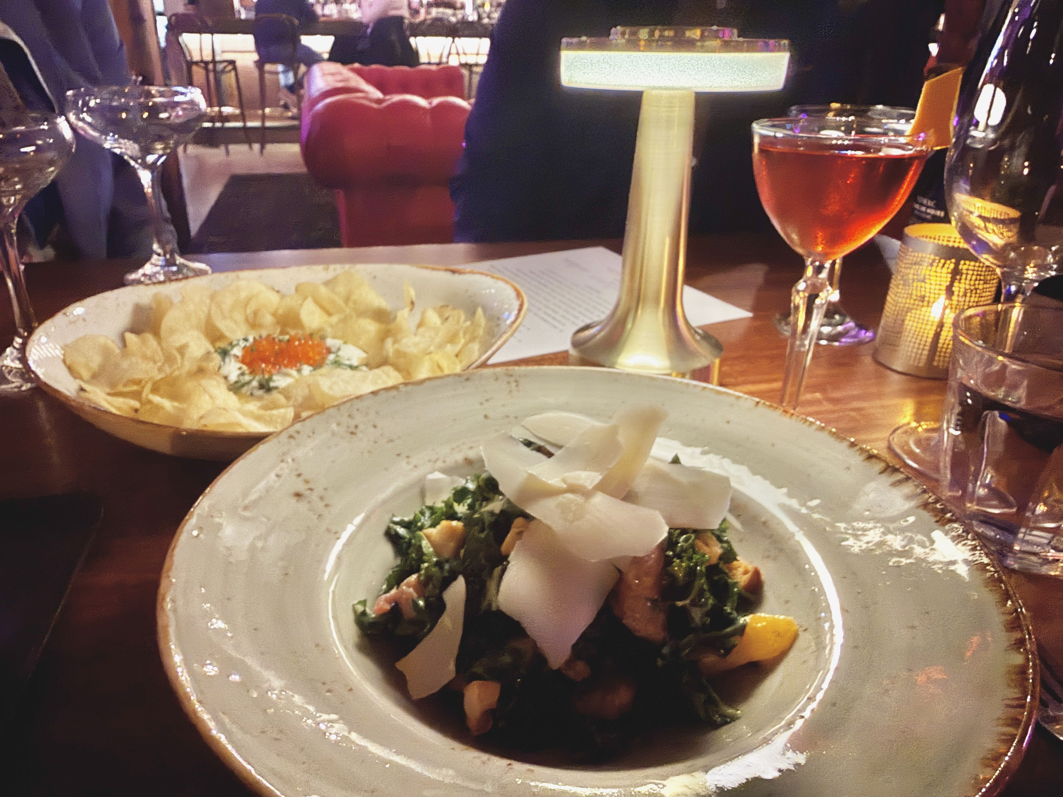 A restaurant table with a dish of greens topped with shaved cheese, a bowl of potato chips with garnish, a pink cocktail, wine glasses, and a warm table lamp in cozy lighting.
