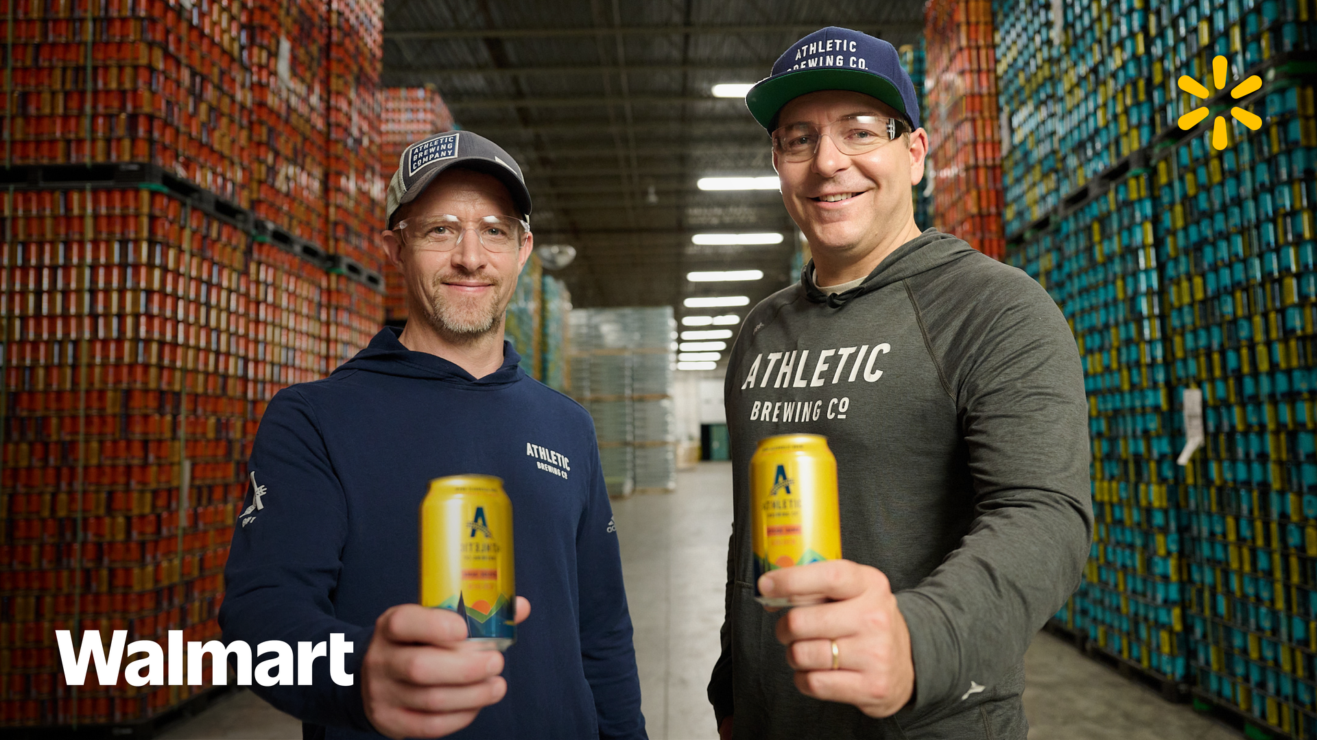 Two smiling individuals hold cans of Athletic Brewing Co. beverages inside a warehouse, with Walmart branding.
