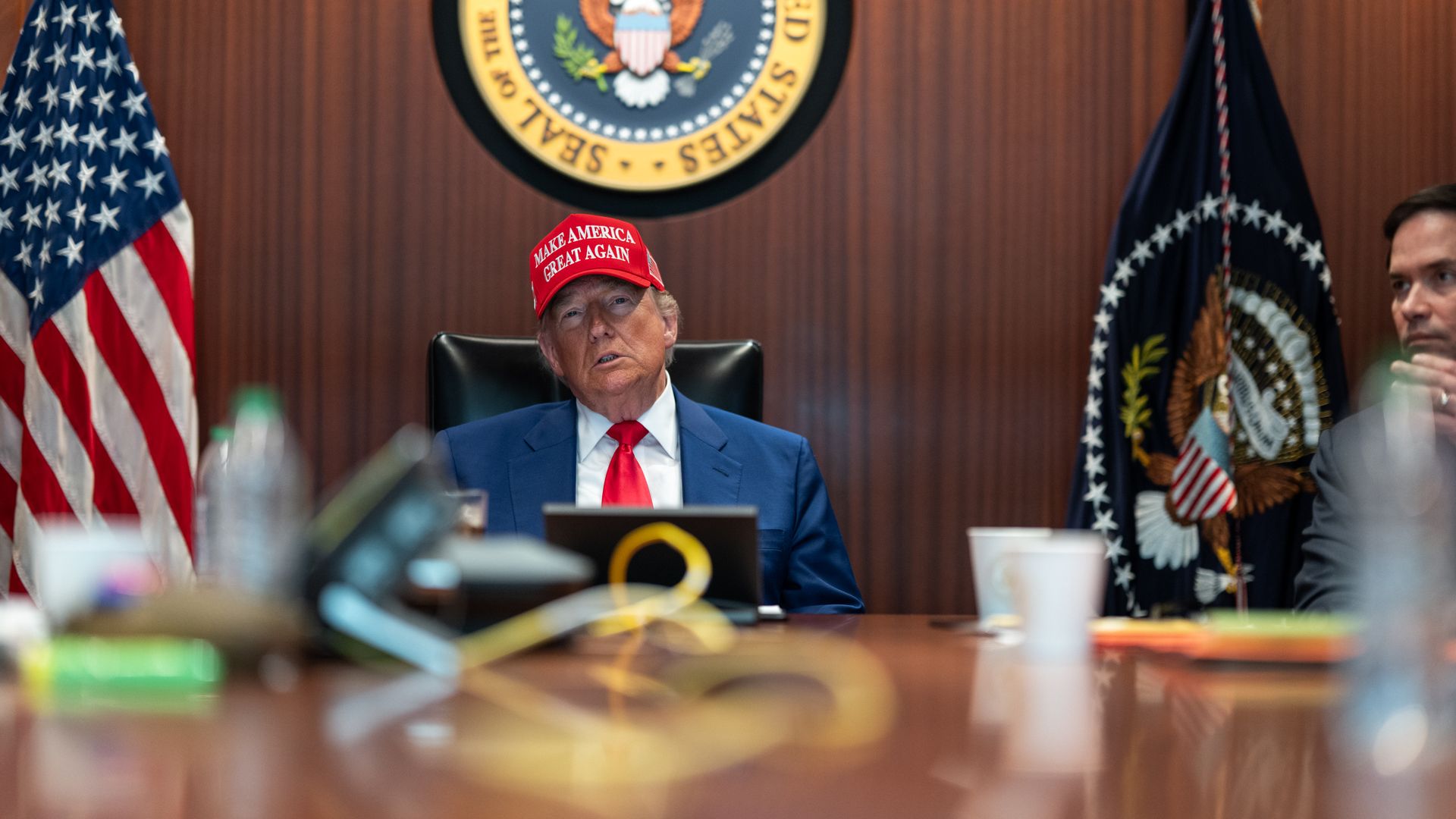 Donald Trump, wearing a blue suit and a red hat and sitting in the wood-paneled situation room.